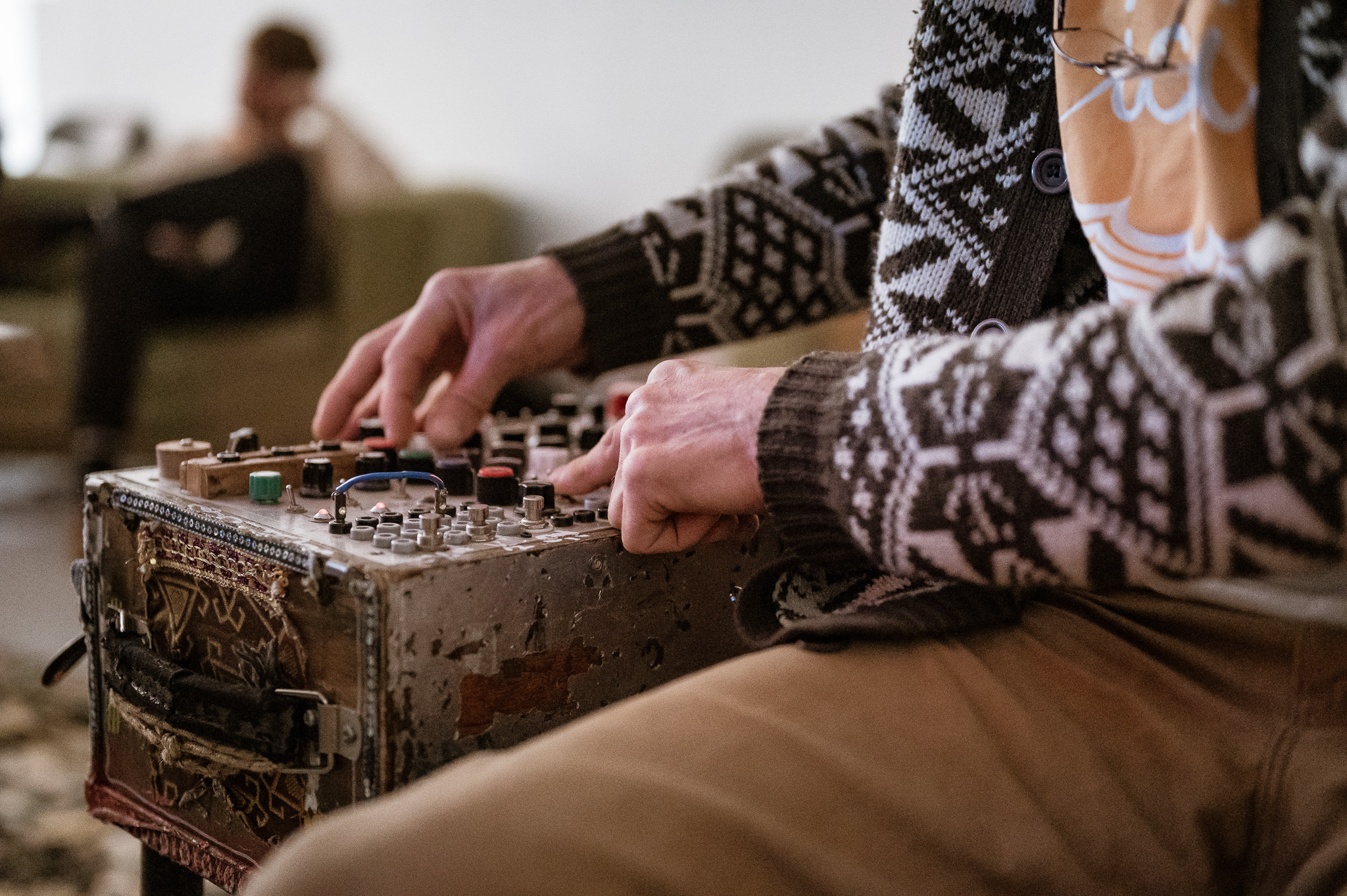 A person wearing a patterned sweater adjusts knobs on a sound device. In the background, someone sits on a sofa.