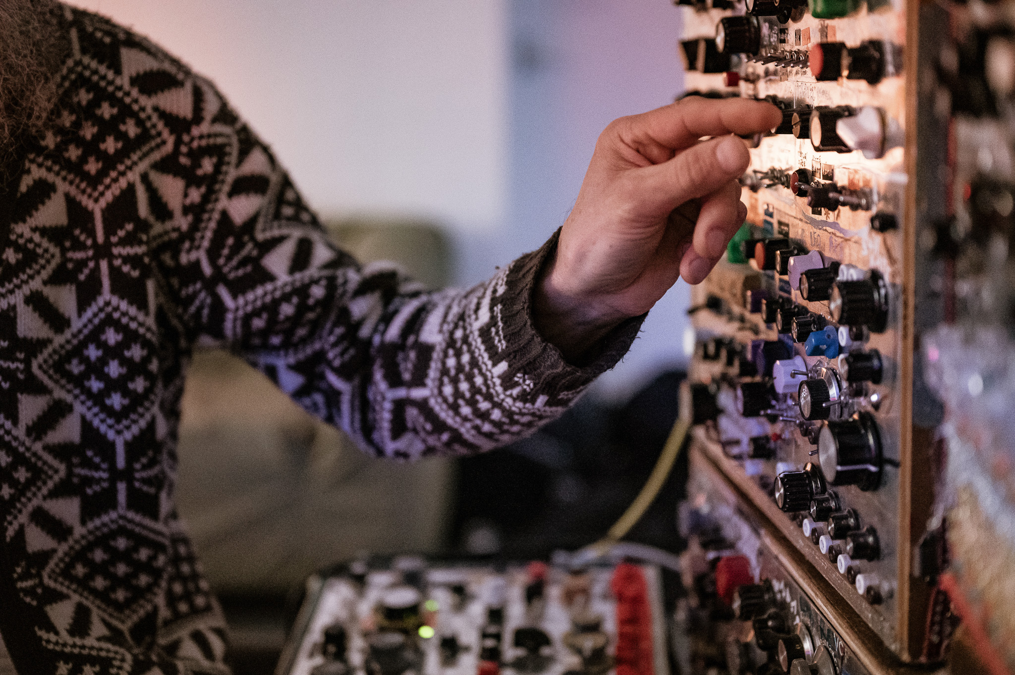 A person wearing a patterned jumper adjusts knobs on a modular synthesiser with various dials, switches, and cables in a softly lit room.