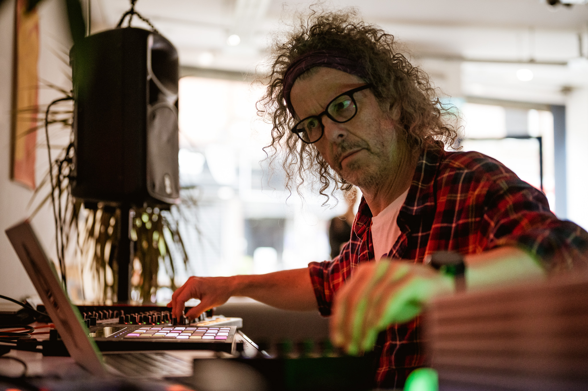 A person with curly hair and glasses, wearing a plaid shirt, focuses intently on operating electronic music equipment in a warmly lit room.