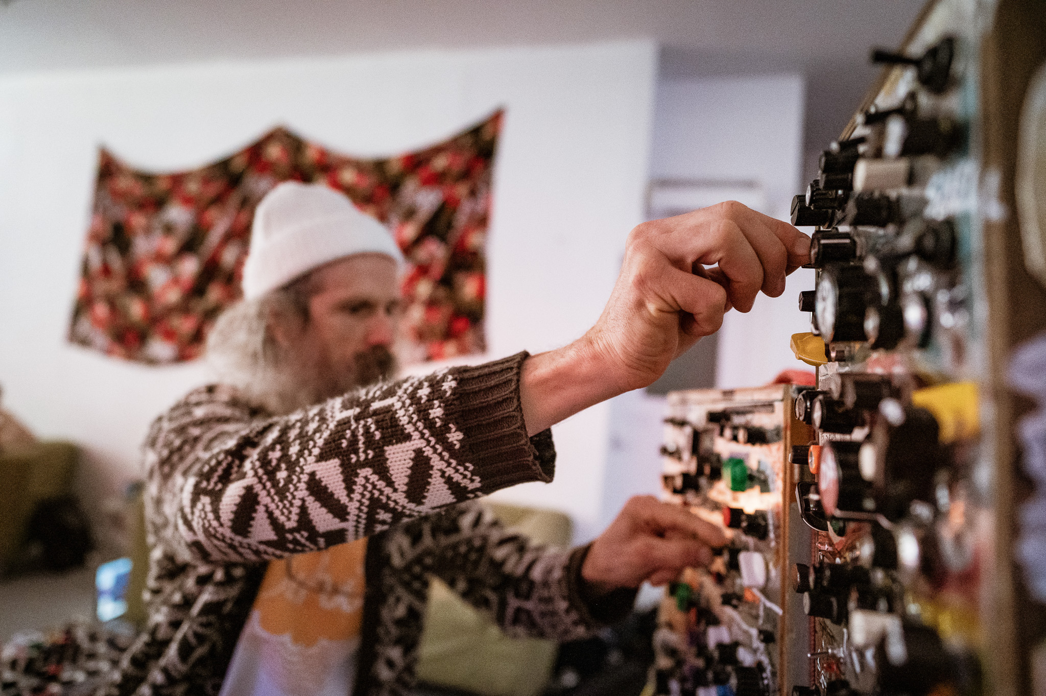 A person with a long beard and a white beanie adjusts knobs on a large modular synthesiser in a cosy room with patterned tapestry on the wall.