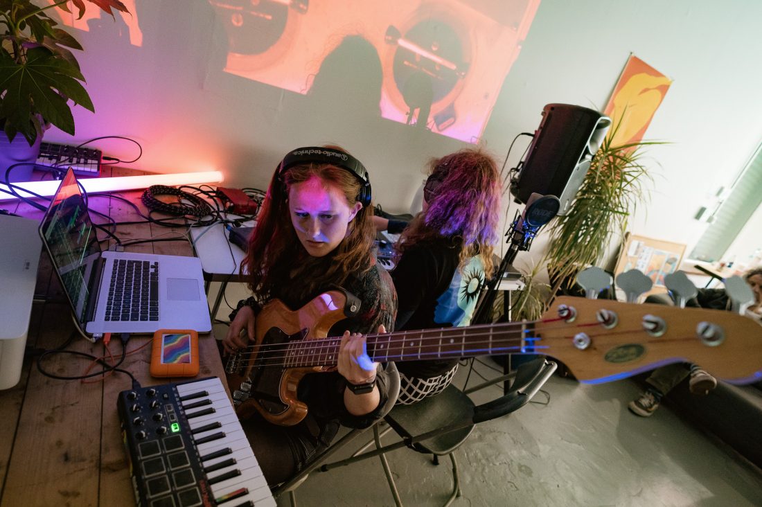 A woman wearing headphones plays an electric bass guitar at a table with a laptop, keyboard, and other audio equipment. Another person operates a music controller. Colourful lights and projections fill the room.