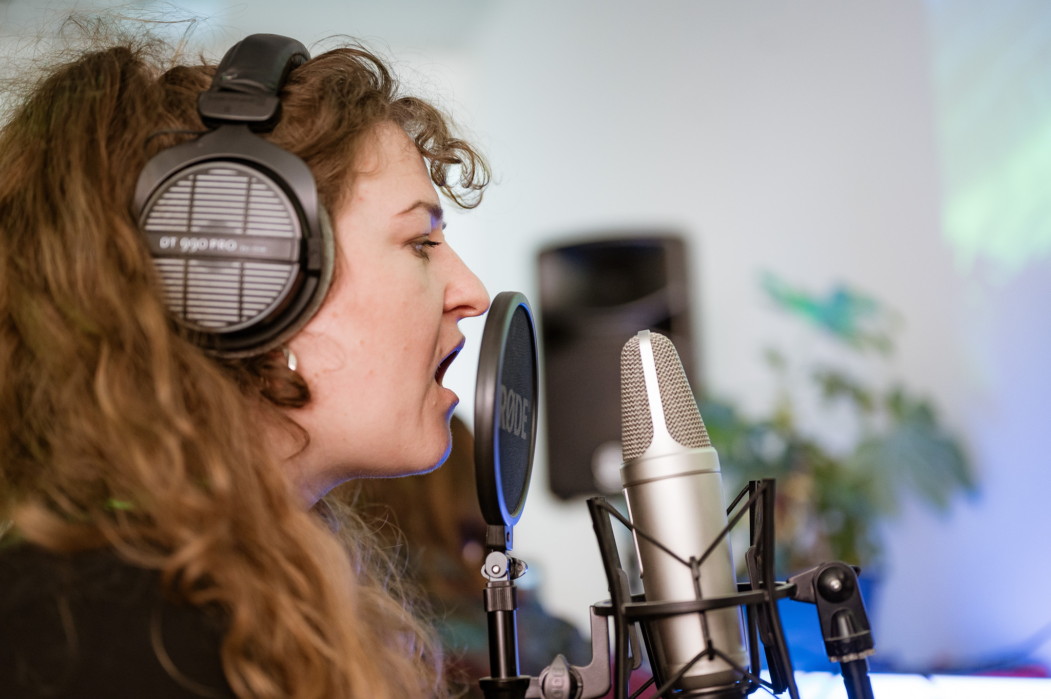 A woman with curly hair wears headphones and sings into a microphone. The setting is a pop up studio, with a blurred speaker and plant in the background.