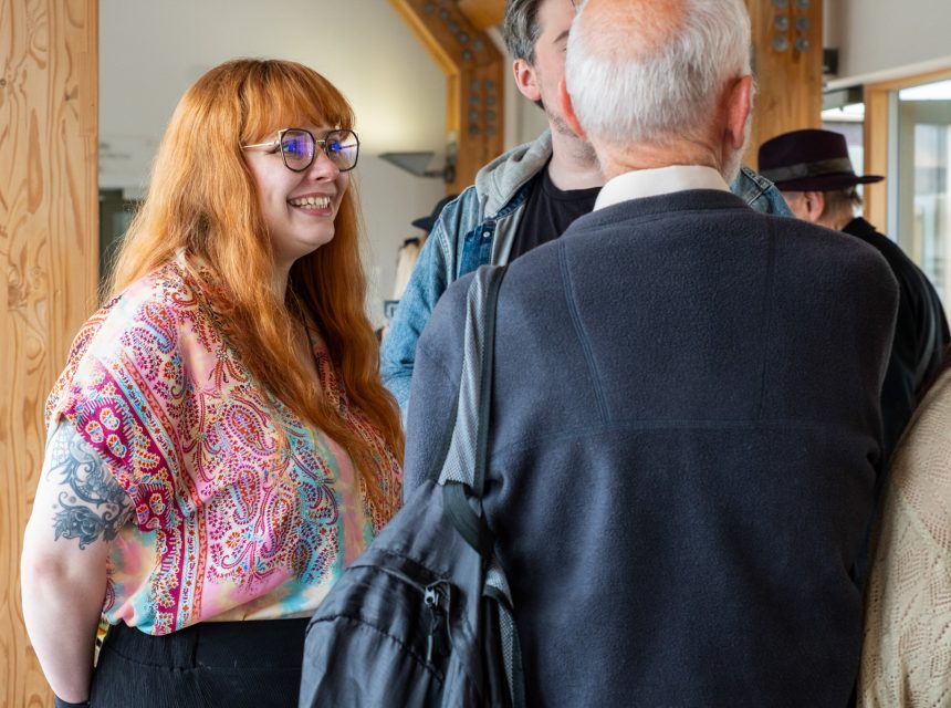 A woman with long red hair and glasses, wearing a colourful patterned shirt, smiles whilst talking to a group of people indoors. The scene appears friendly and social.