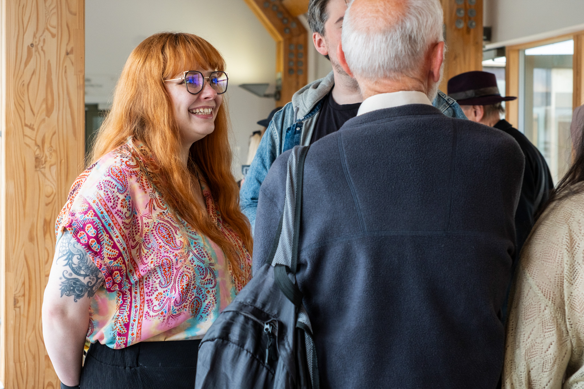 A woman with long red hair and glasses, wearing a colourful patterned shirt, smiles whilst talking to a group of people indoors. The scene appears friendly and social.