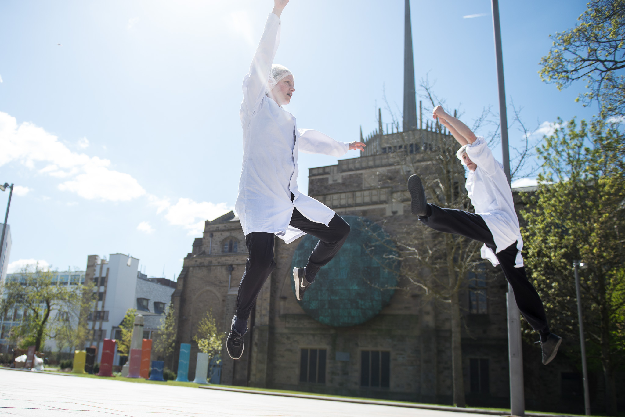 Two people in white coats and black trousers leap into the air with arms raised, performing dynamic jumps outside on a sunny day, with a large building featuring a spire in the background.