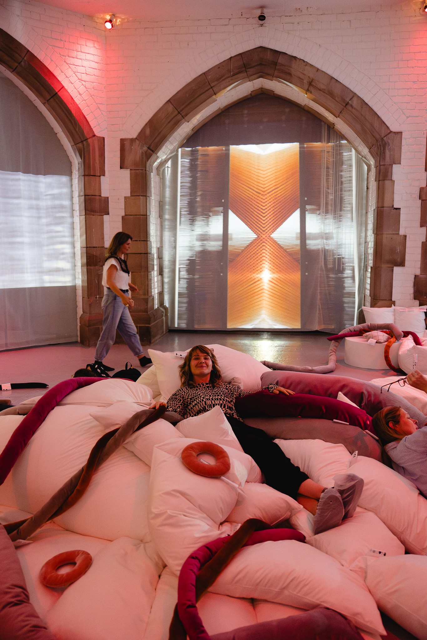A woman relaxes on oversized white and pink doughnut-shaped cushions as part of an art installation. Behind her, another person walks past arched doorways where a large screen displays abstract images of machinery.