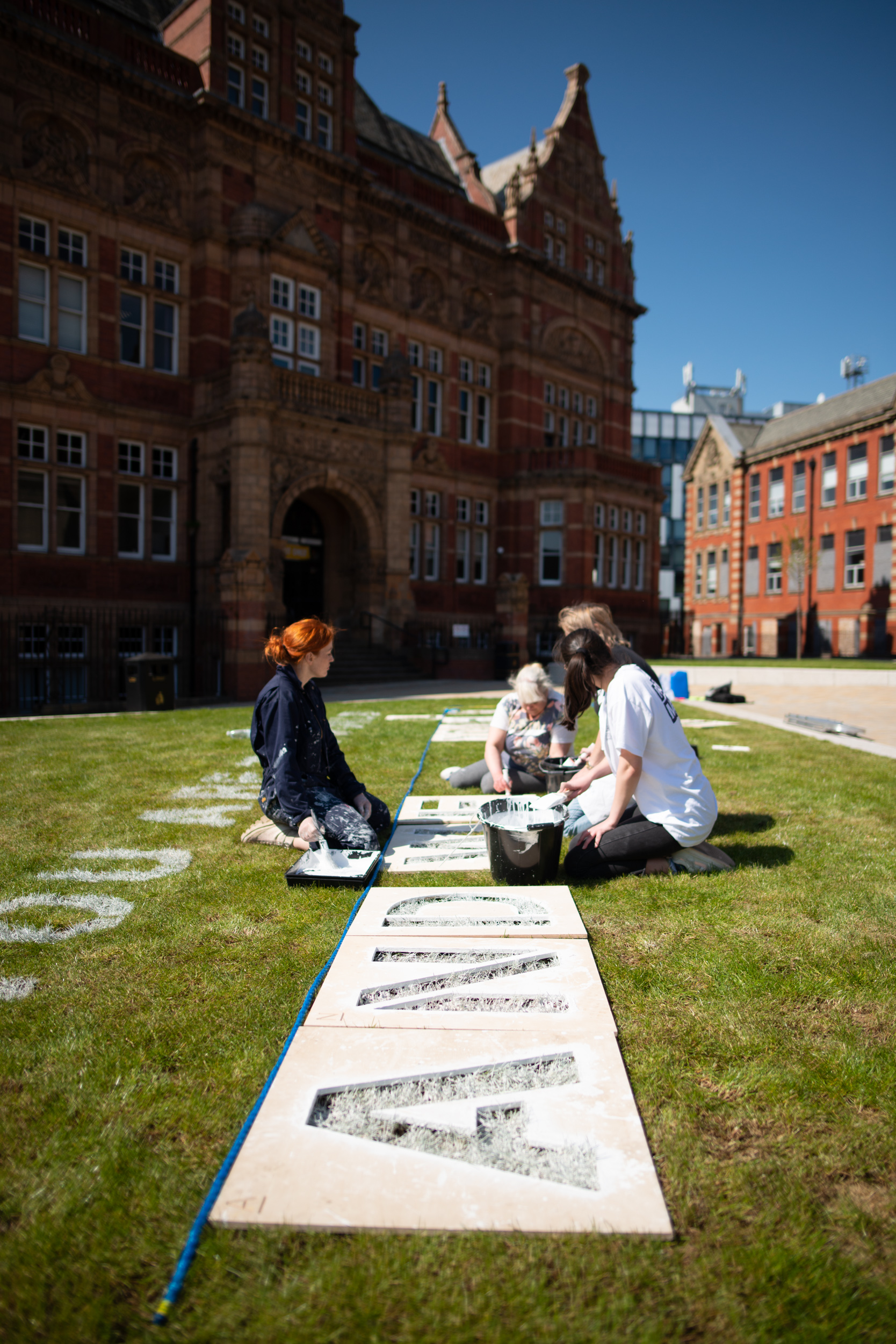 Three people paint large stencilled letters on white paper laid out on grass in front of a historic red brick building under a clear blue sky.