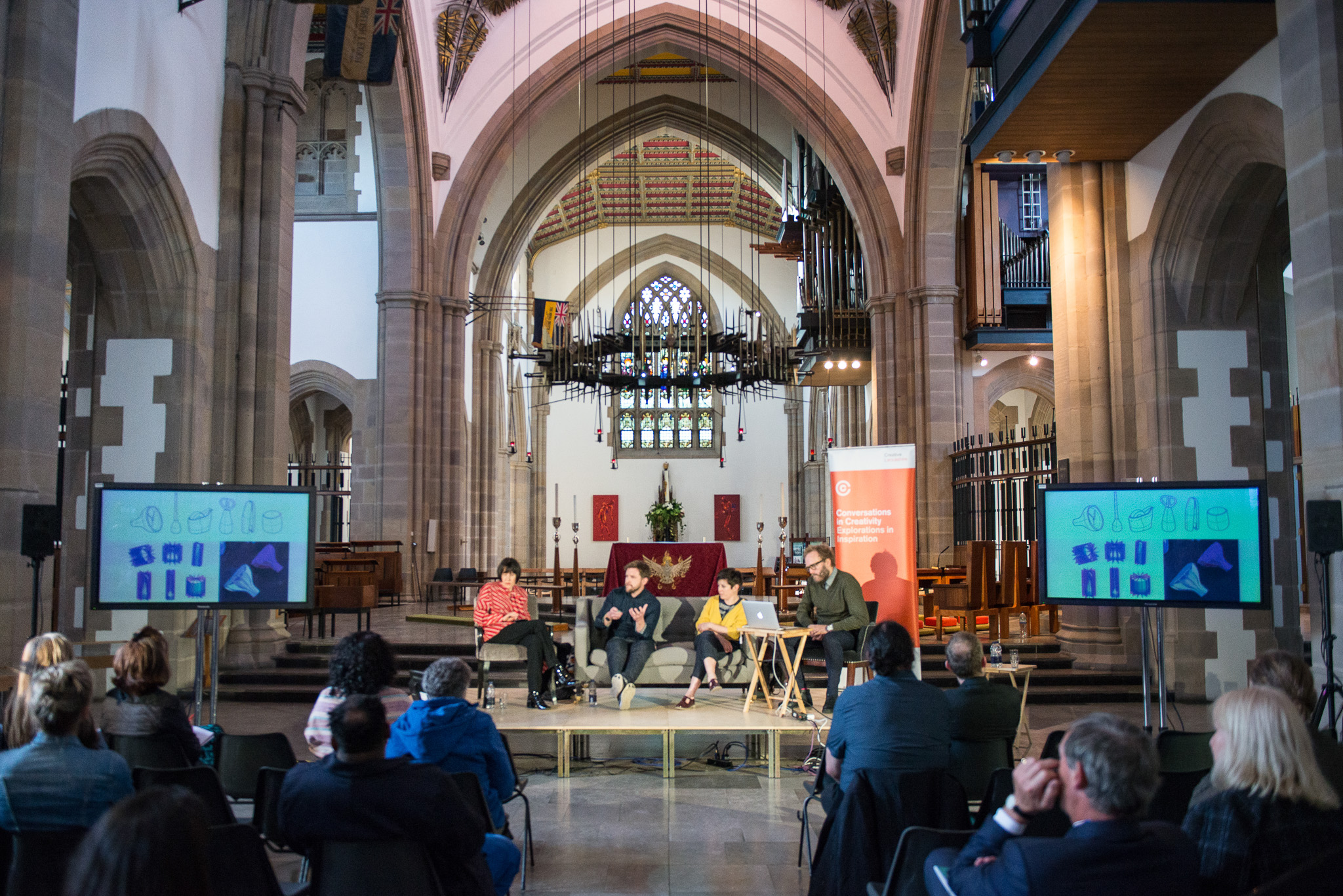 A panel of five people sits on a stage in a church with stained glass windows, speaking to an audience. Two screens display images on either side, and a banner stands near the panel. The setting is bright and spacious.