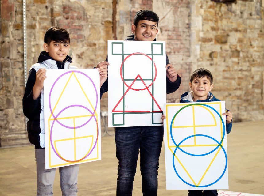 Three boys stand indoors in front of a brick wall, each smiling and holding a large poster featuring colourful geometric shapes, including circles, triangles, and squares.