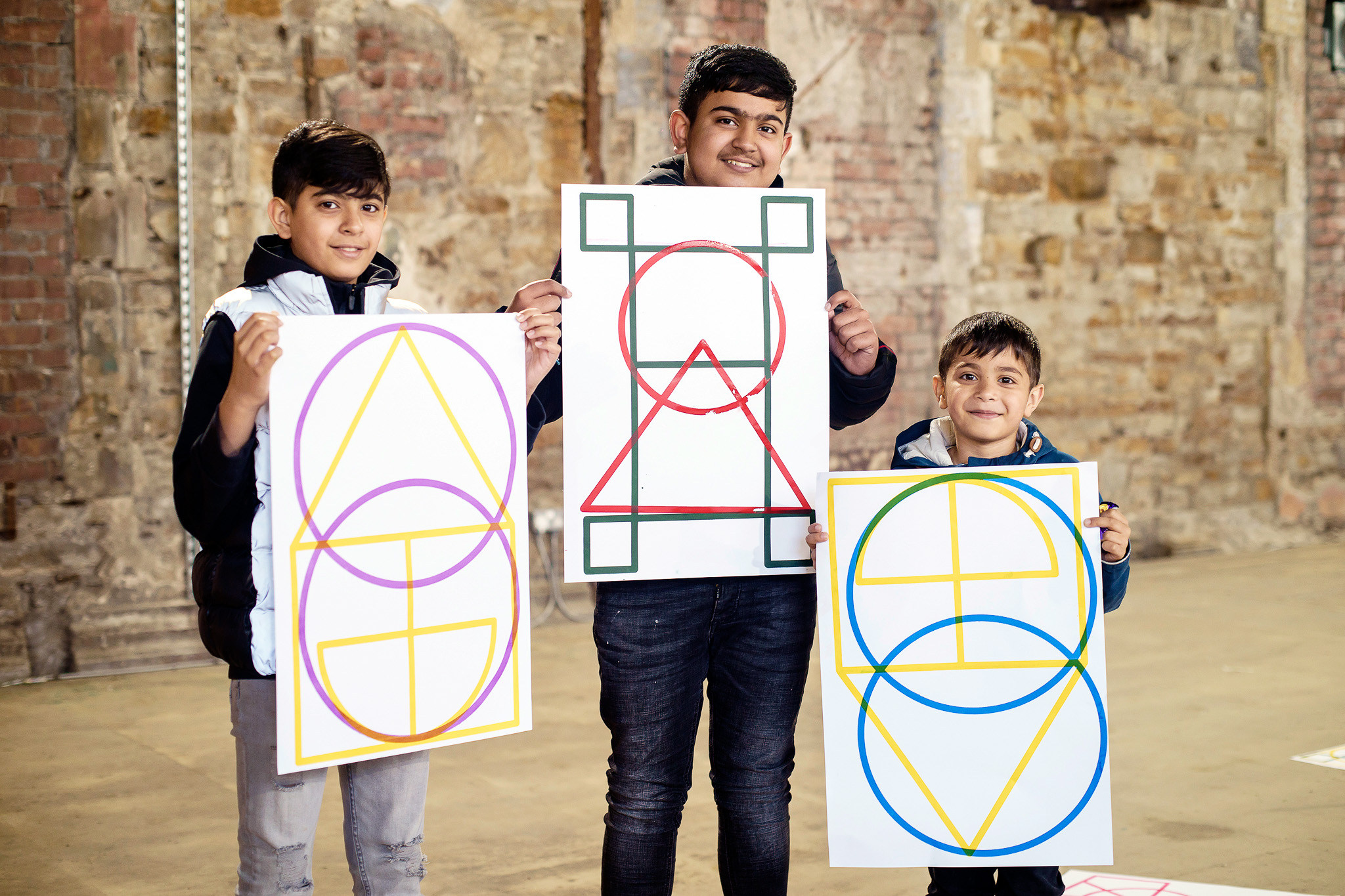 Three boys stand indoors in front of a brick wall, each smiling and holding a large poster featuring colourful geometric shapes, including circles, triangles, and squares.