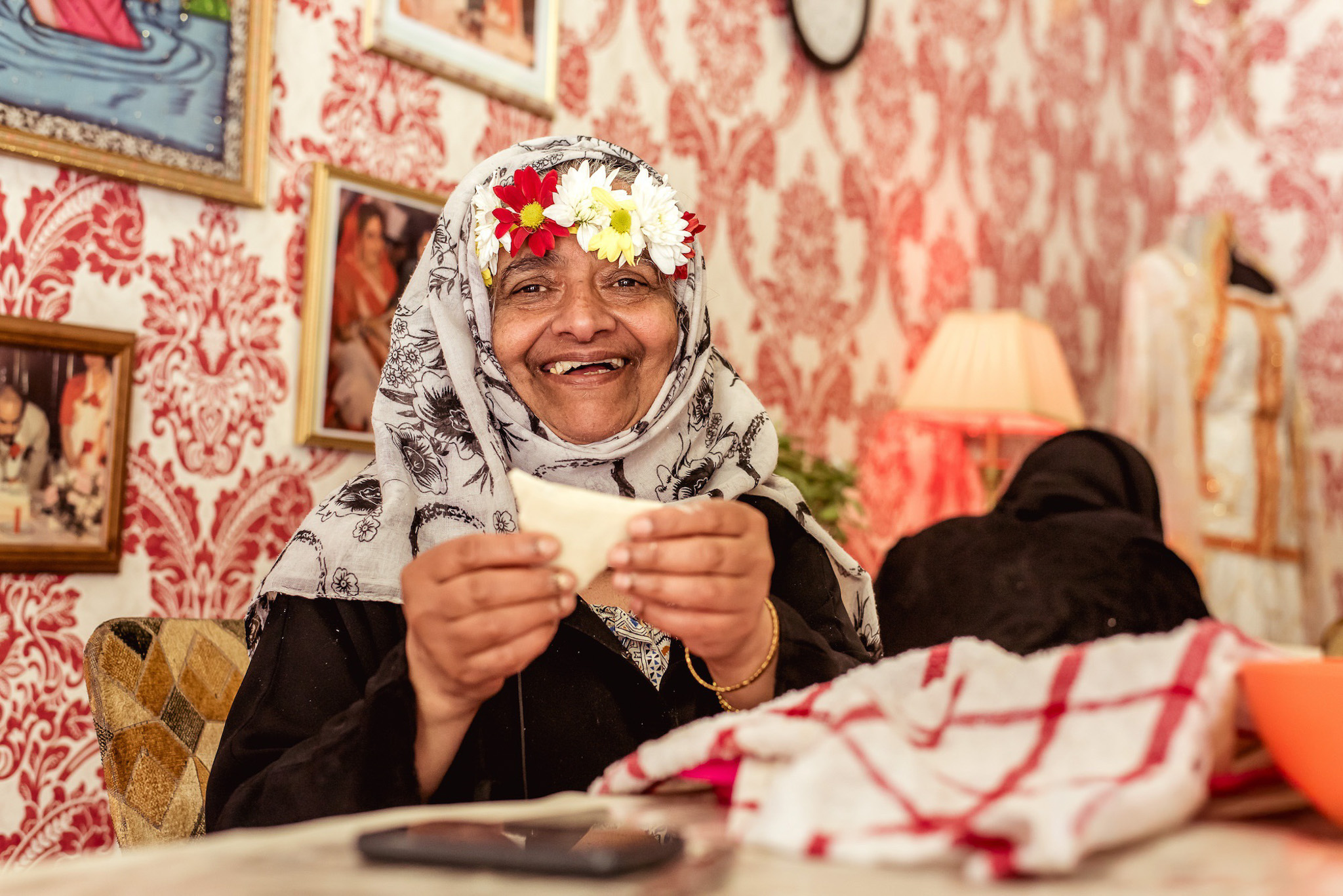 An older woman wearing a floral headscarf and flower crown smiles whilst holding dough in her hands at a table. The room has red patterned wallpaper, framed photos, and a cosy atmosphere.