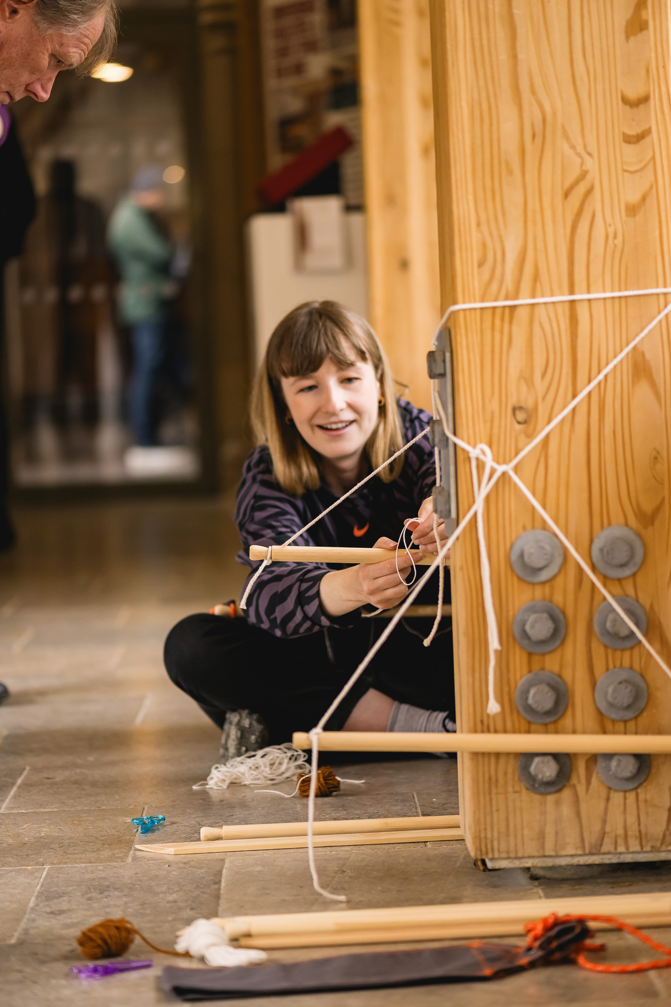 A young woman crouches on the floor, smiling as she weaves white string through a large wooden structure indoors. Various yarns and rods are scattered around her. Another person stands nearby.