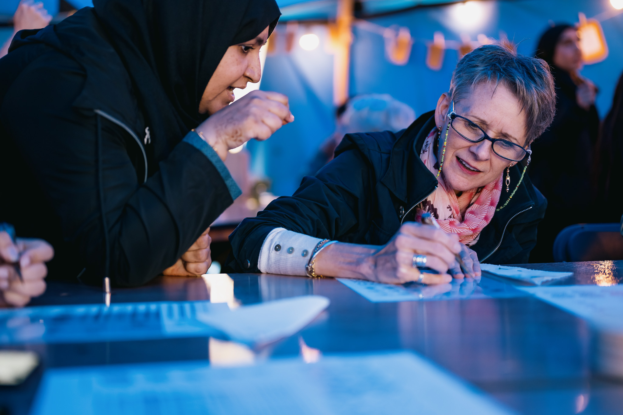 Two women sit at a table, one wearing a hijab and the other glasses and a scarf. They are engaged in an activity, writing or drawing on paper, under blue lighting at an event.