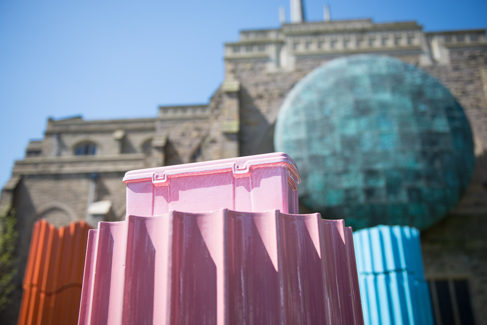 A close-up of a pink, fluted sculpture resembling an ice lolly, with similar blue and orange sculptures nearby. In the background stands a historic stone building with a large, oxidised copper dome under a clear sky.
