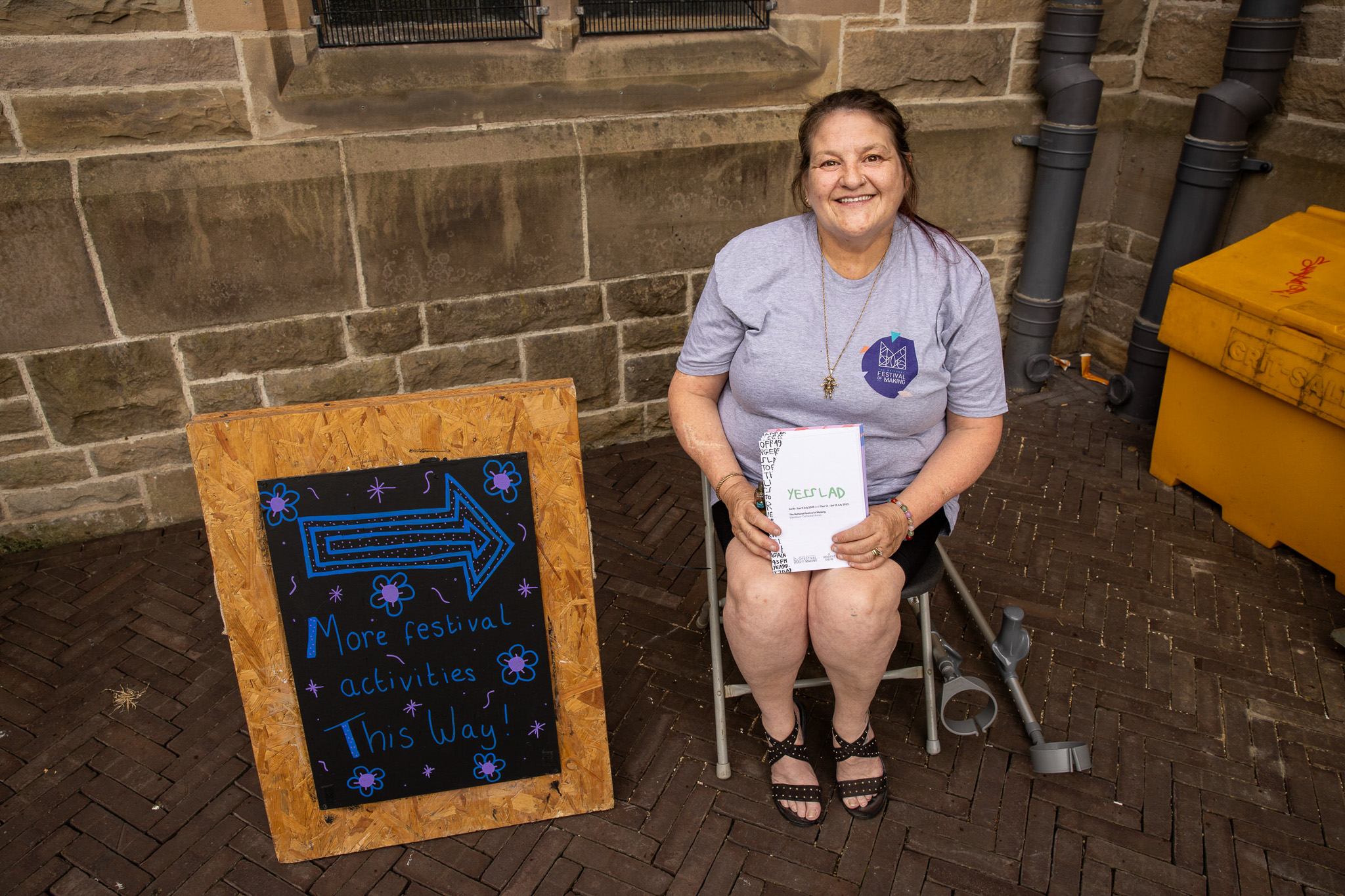 A smiling woman sits on a chair near a stone wall, holding papers. A crutch rests beside her. Next to her is a sign with a blue arrow and text reading, “More festival activities this way!” decorated with flowers.