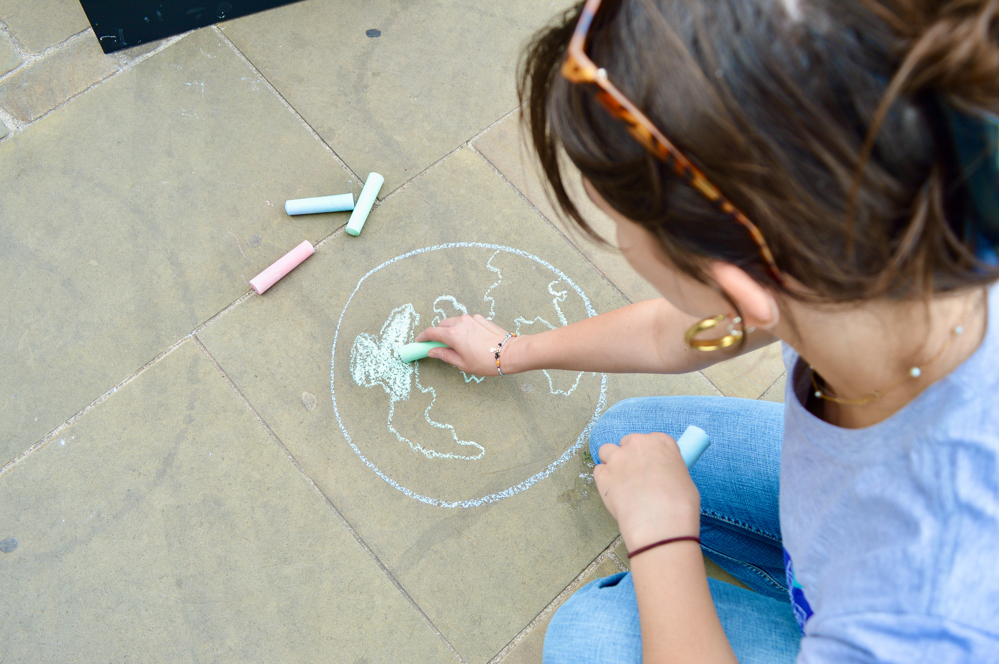 A person draws a chalk outline of the Earth on pavement, colouring in continents with green chalk. Other pieces of coloured chalk lie nearby. The person wears sunglasses on their head and large hoop earrings.