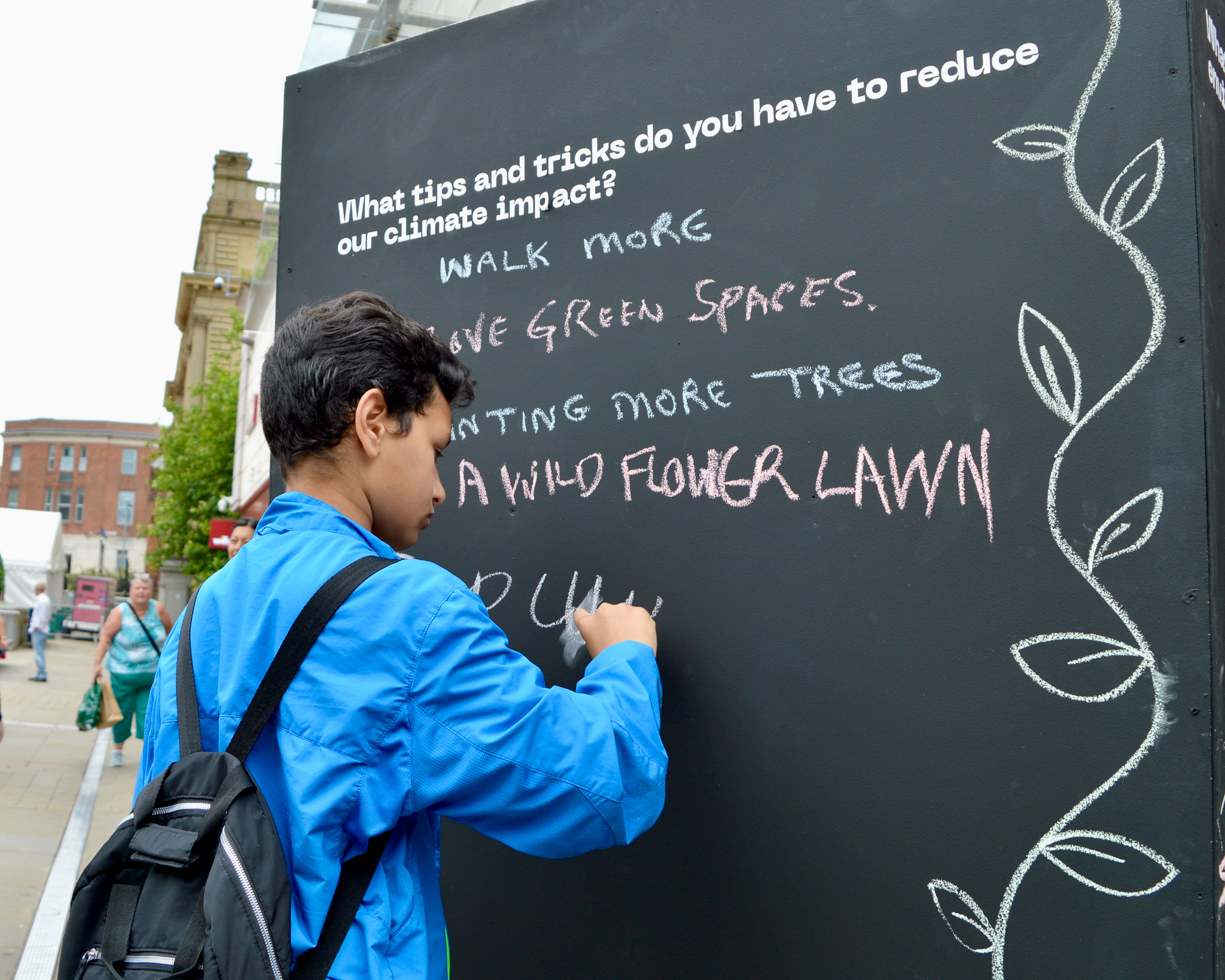 Person in blue jacket writes climate tips on a chalkboard in a public space, suggesting ways to reduce environmental impact.