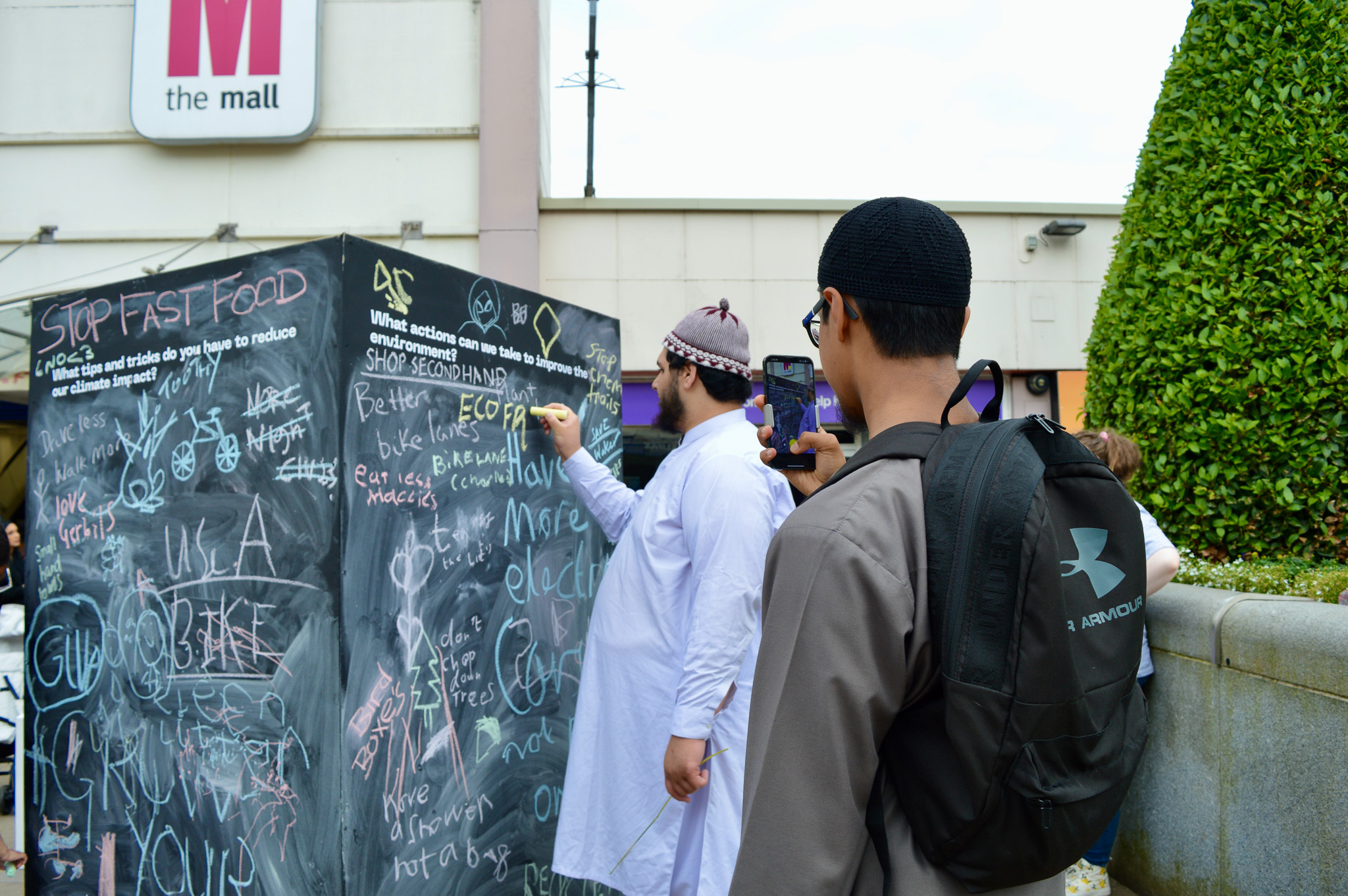 Two men in traditional clothing write messages on a large outdoor chalkboard filled with colourful chalk writing. One man films the scene on his phone; a shopping centre sign and greenery are visible in the background.