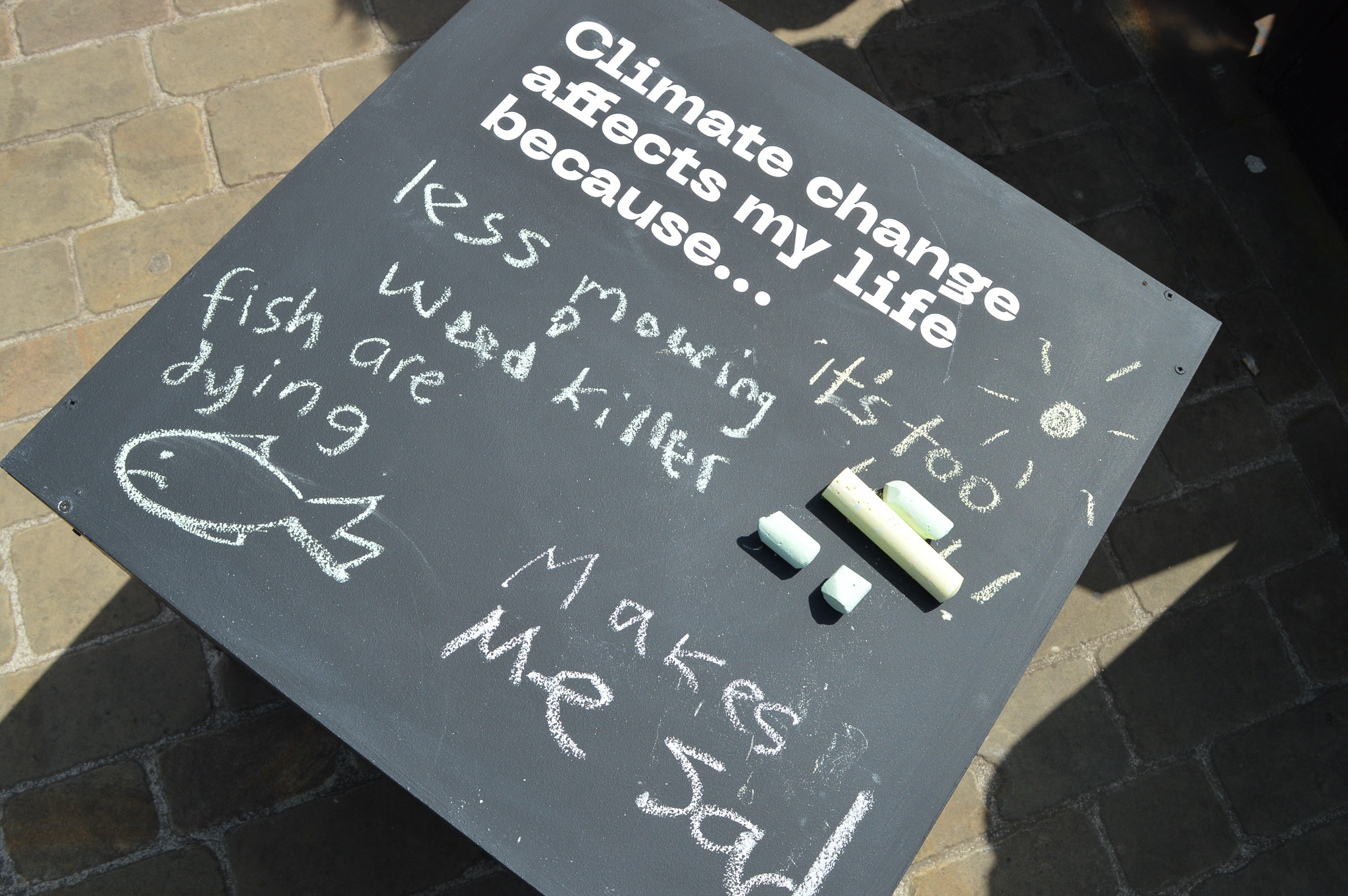 A blackboard on the ground reads “Climate change affects my life because…” with chalk responses like “fish are dying,” “less mowing,” “weed killer,” “it’s too hot” with a sun drawing, and “makes me sad”; chalk pieces are on the board.