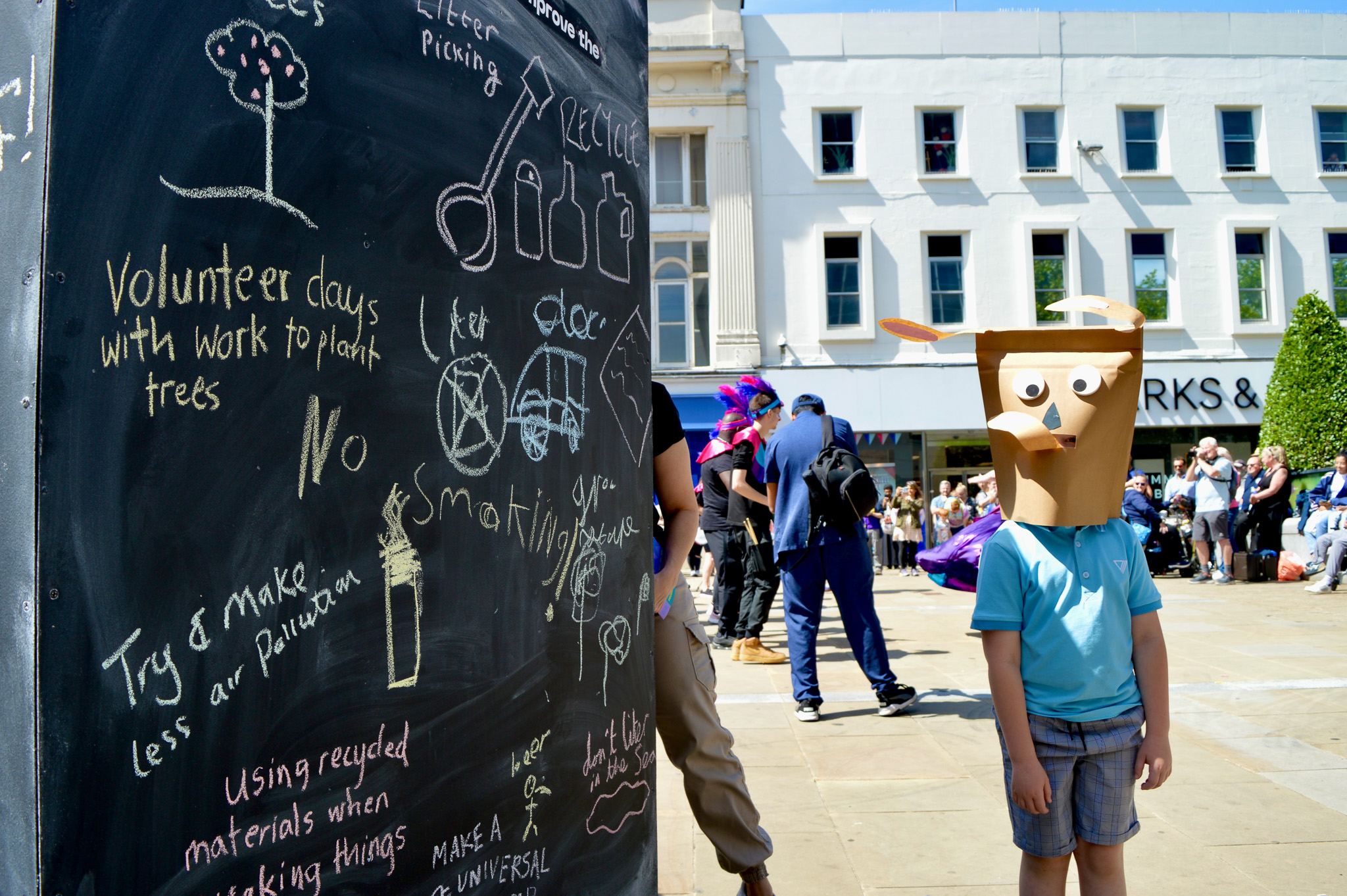 A child wearing a cardboard animal mask stands next to a large blackboard covered in drawings and messages about recycling and volunteering, in a busy outdoor square with people gathered in the background.