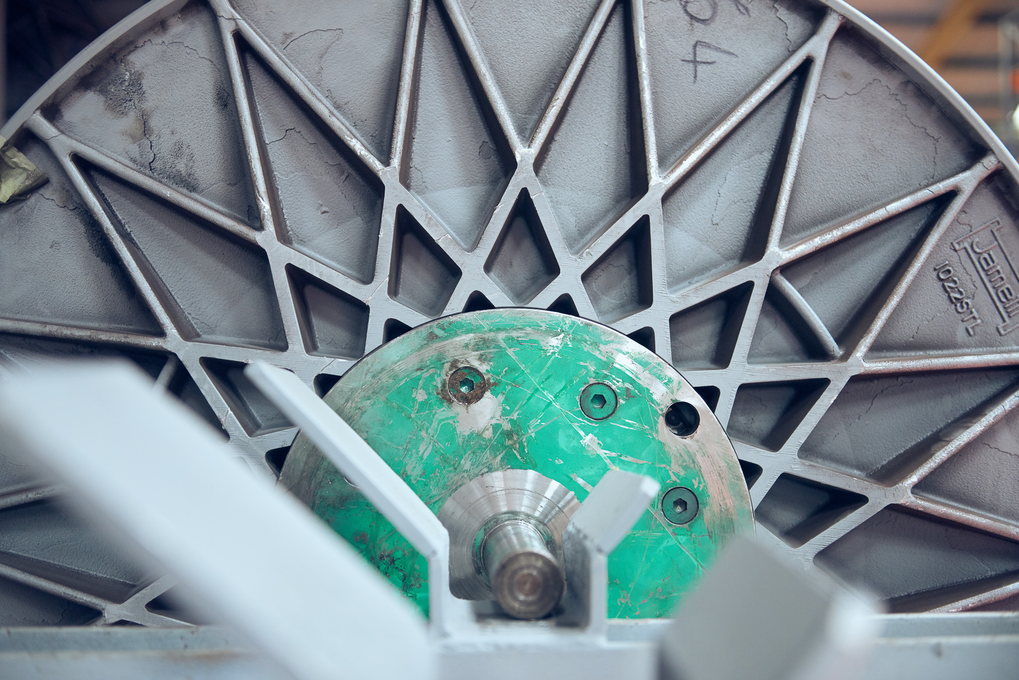 Close-up of a large industrial machine part with a geometric, star-like metal pattern and a central green circular component, showing bolts and metallic textures.