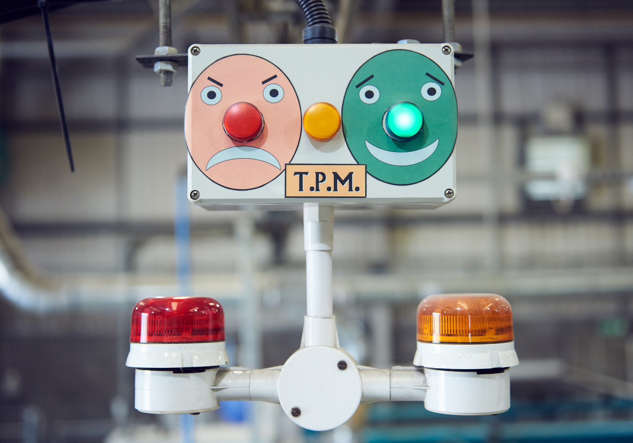 A control panel with a red angry face and a green happy face, each with a corresponding light. The green light is lit. Two warning beacons, one red and one orange, are attached below the panel labelled “T.P.M.”.