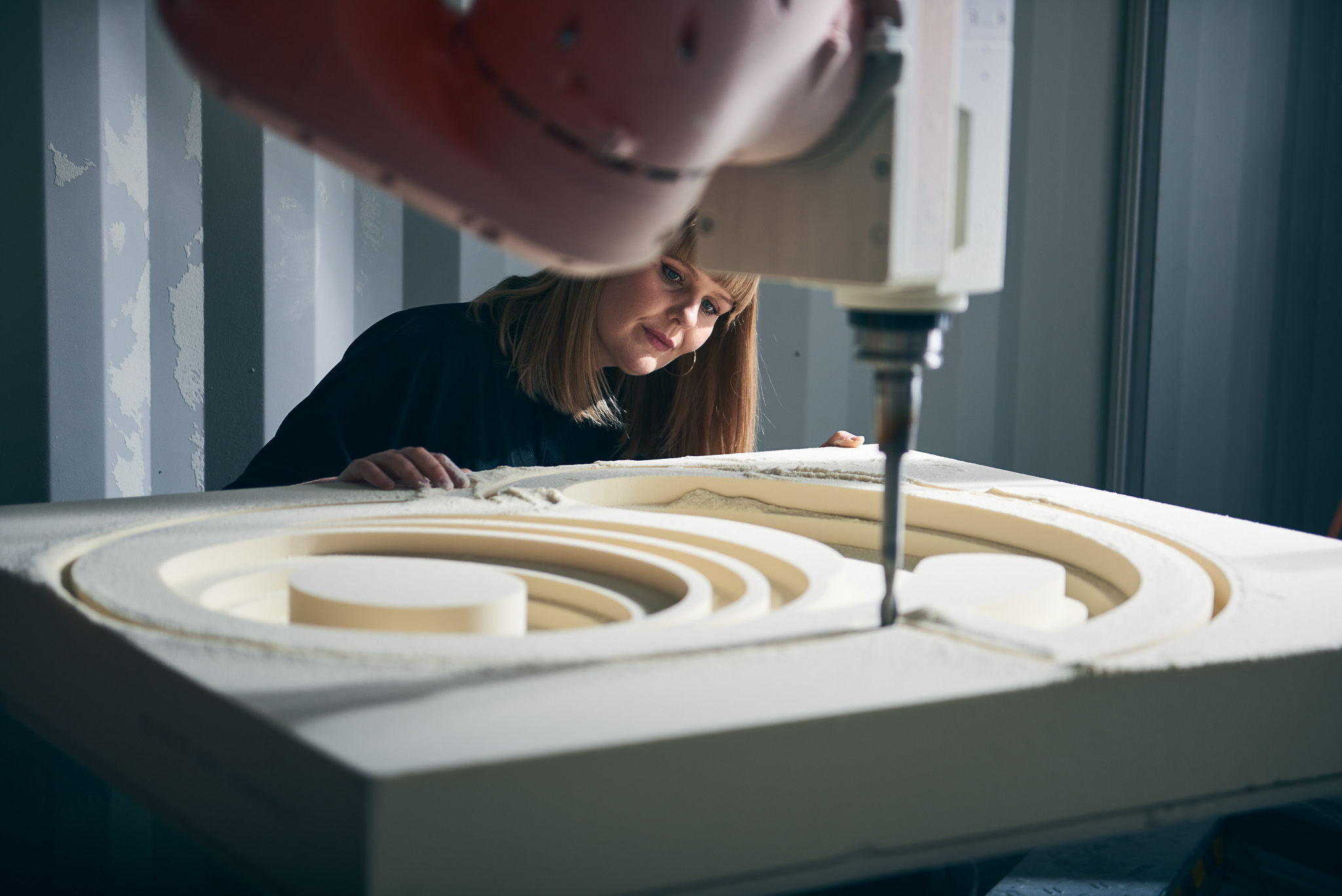 Liz Wilson observes a large CNC machine carving circular patterns into a thick piece of foam in an industrial workshop setting.