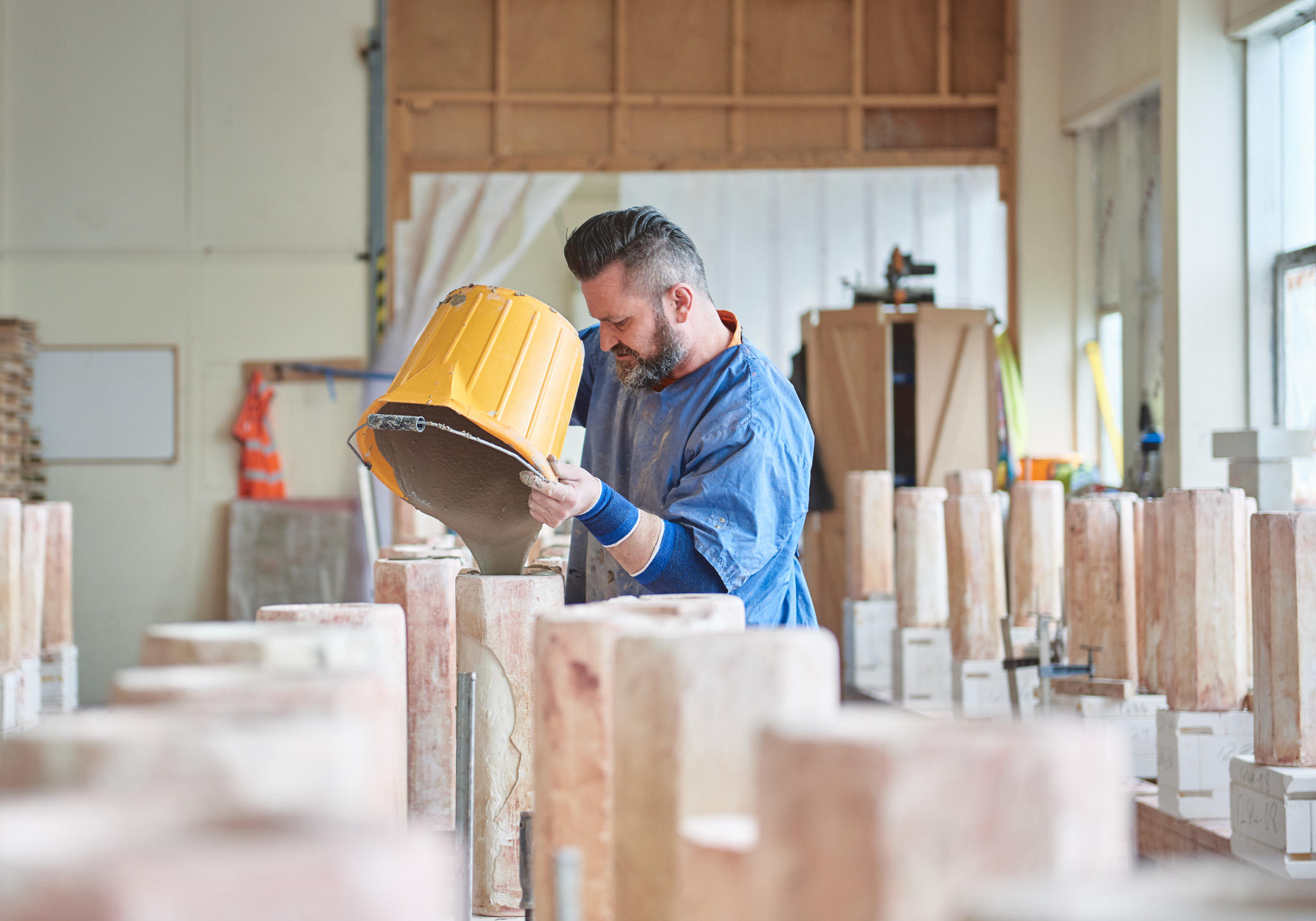 A man wearing a blue shirt pours liquid clay from a large yellow container into a mould at a workshop filled with rows of upright, rectangular moulds. Sunlight streams in through windows on the right.