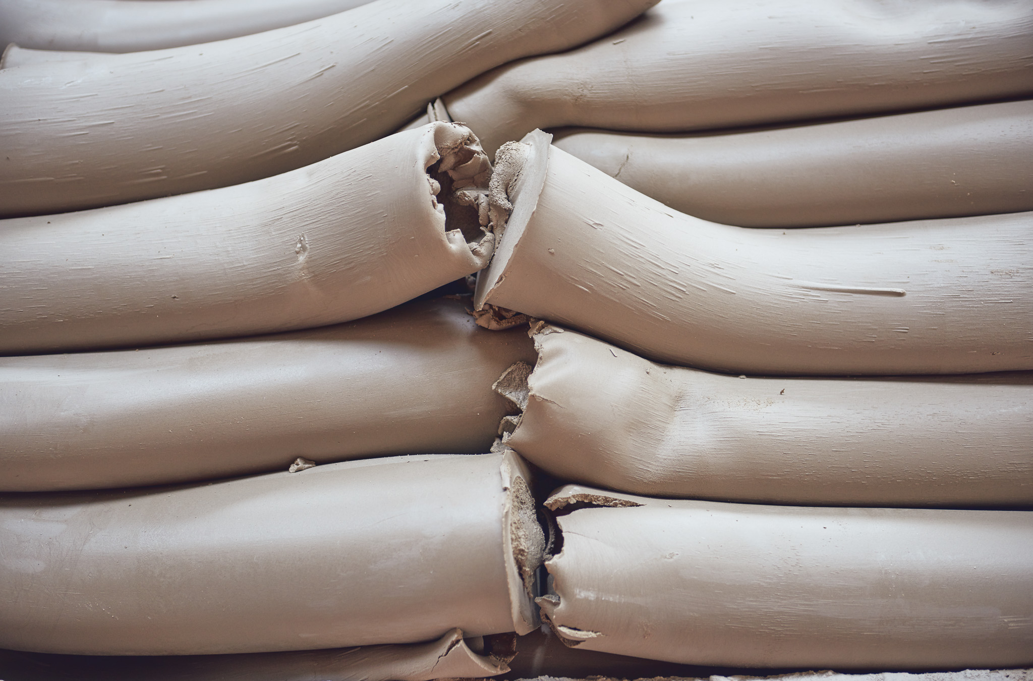 Several thick, smooth, beige tubes of clay are stacked horizontally, with some tubes slightly bent or compressed where they meet, showing textured and rougher ends.