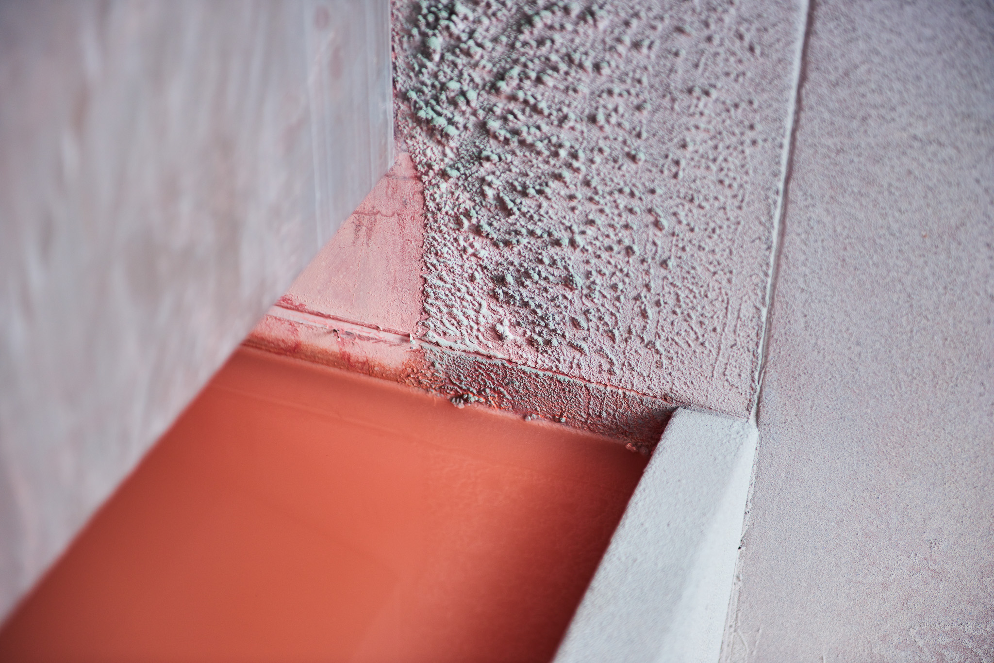 Close-up of a textured wall corner painted in shades of pink and white, with a smooth, flat surface below in a reddish-orange colour. The image highlights contrasts between rough and smooth surfaces.