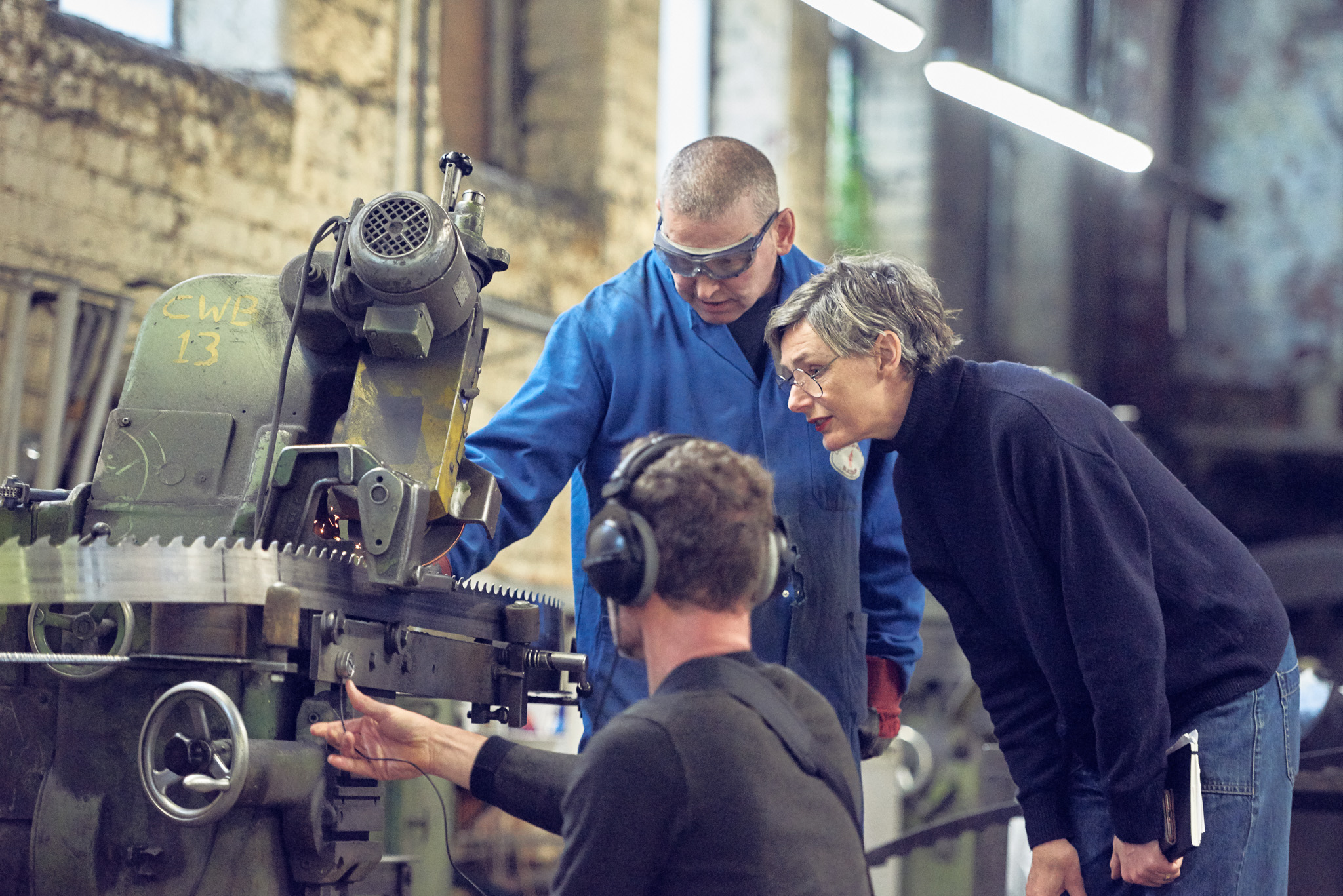 Three people in a workshop examine a large industrial machine. One person wears blue overalls and safety goggles, another leans in holding a notepad, and the third, wearing headphones, operates the machine.