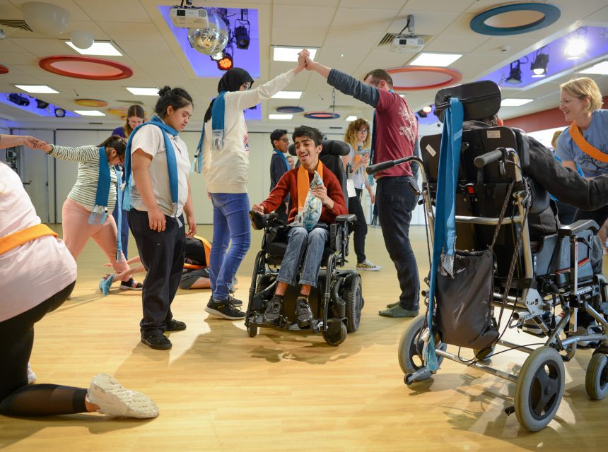 A group of people, some using wheelchairs, dance and interact with support staff in a brightly lit room with colourful ceiling decorations. Two people high-five in the centre, smiling and enjoying the activity.