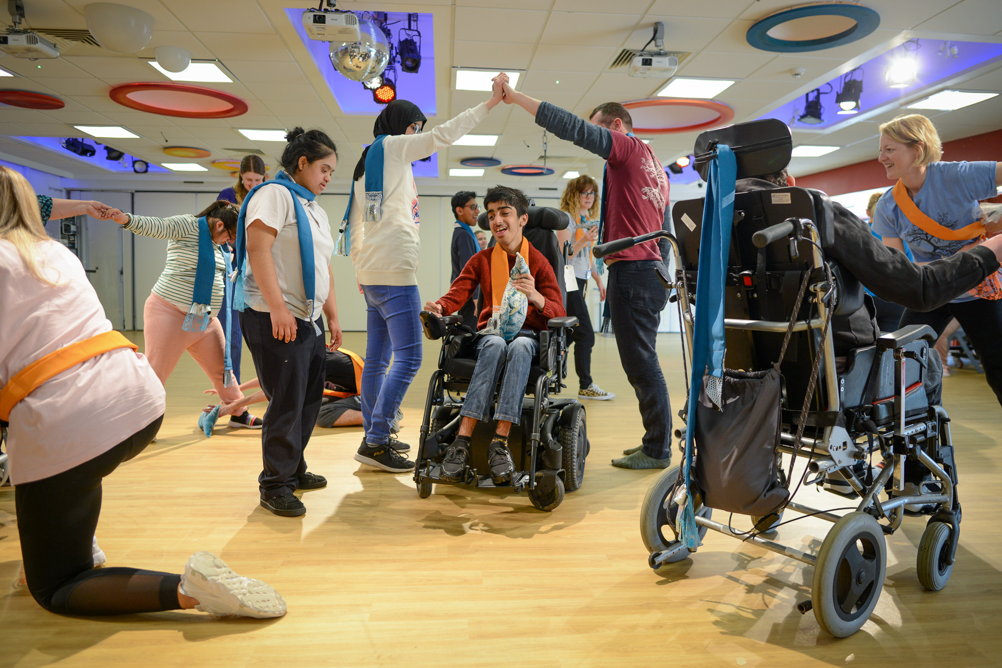 A group of people, some using wheelchairs, dance and interact with support staff in a brightly lit room with colourful ceiling decorations. Two people high-five in the centre, smiling and enjoying the activity.