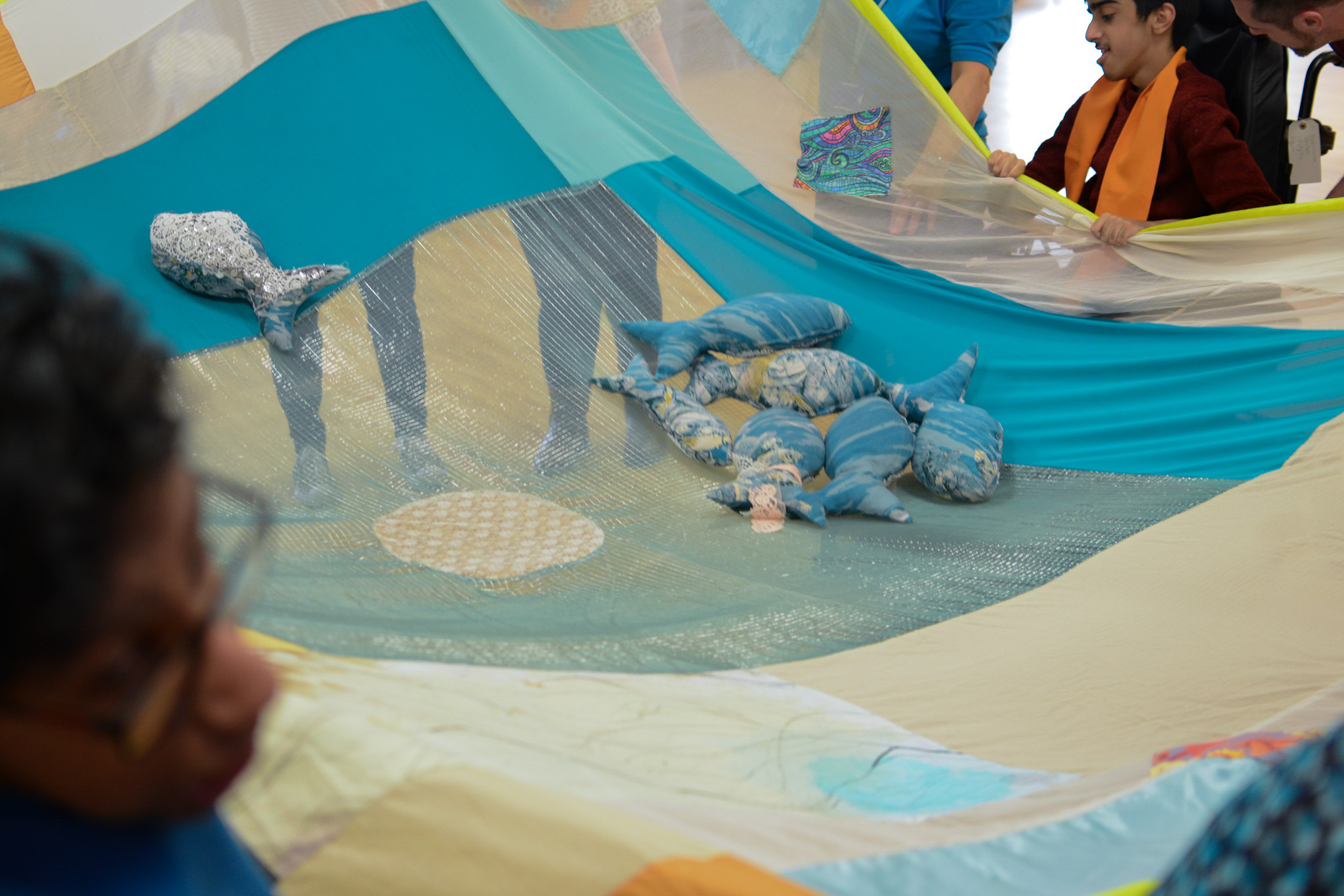 Children and adults play with a large, colourful fabric parachute, decorated with sea-themed soft toys like dolphins and a whale. The parachute is being lifted, creating waves and motion in the fabric.