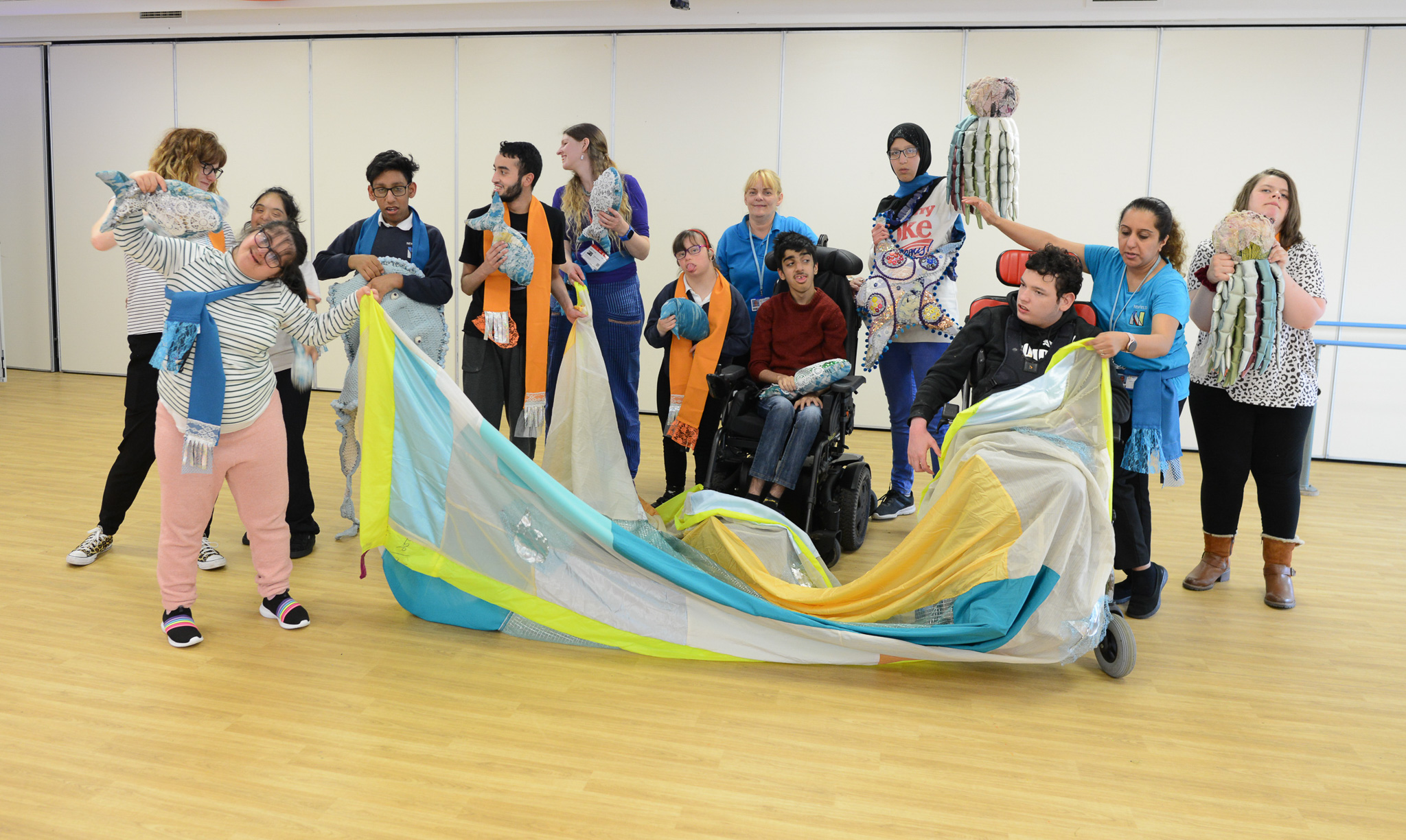 A diverse group of people, including a person in a wheelchair, are holding colourful scarves and fabrics, smiling and interacting together in a bright room with a wooden floor.