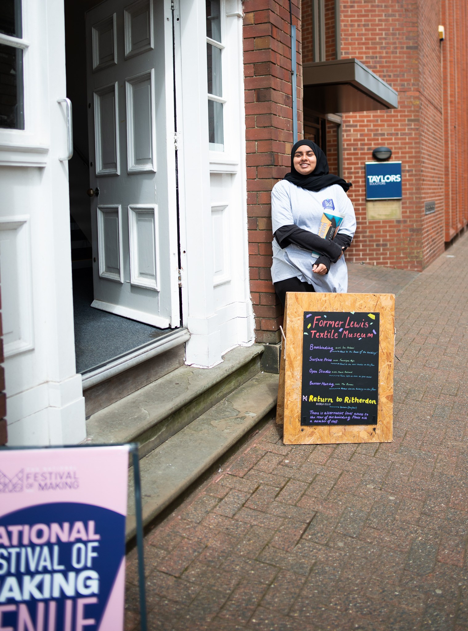 A woman in a hijab and white shirt stands smiling by an open doorway next to a sign listing events for the Former Louis Textile venue during the National Festival of Making. Brick buildings line the walkway.