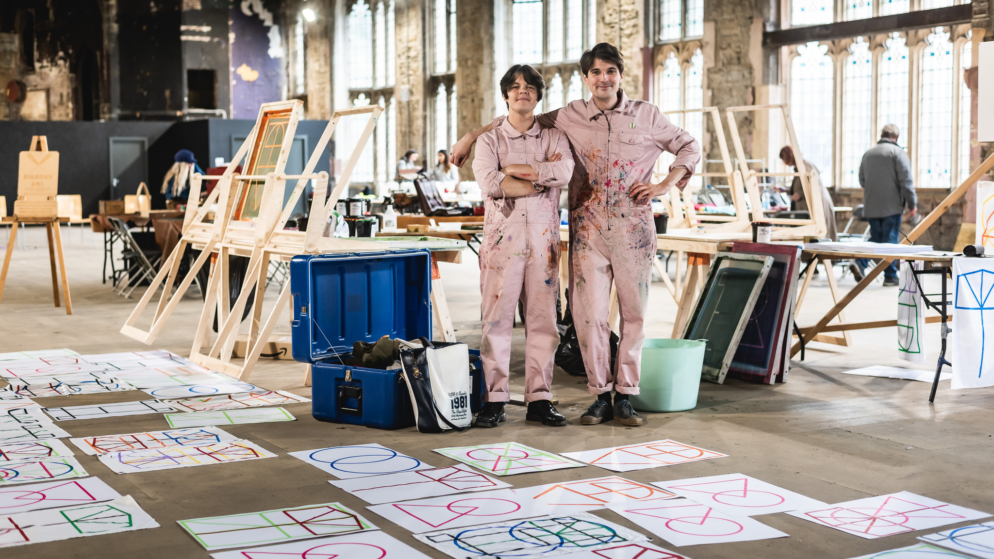 Two people wearing paint-splattered overalls stand in a large, sunlit hall surrounded by printed geometric artwork on the floor, screen printing materials, and various art supplies.