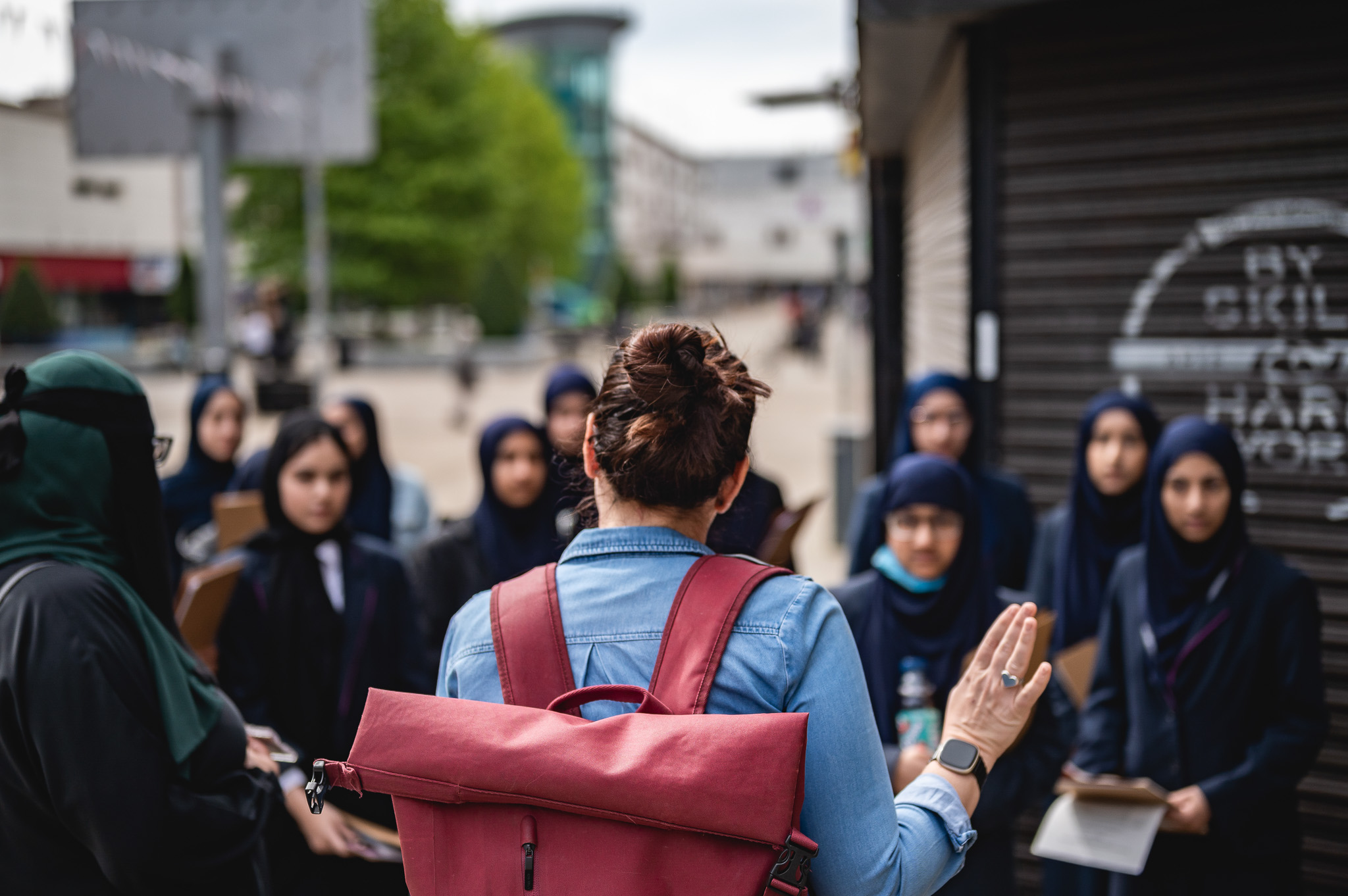 A woman with a red rucksack addresses a group of students in dark clothing and headscarves outdoors, with urban buildings and trees in the background.