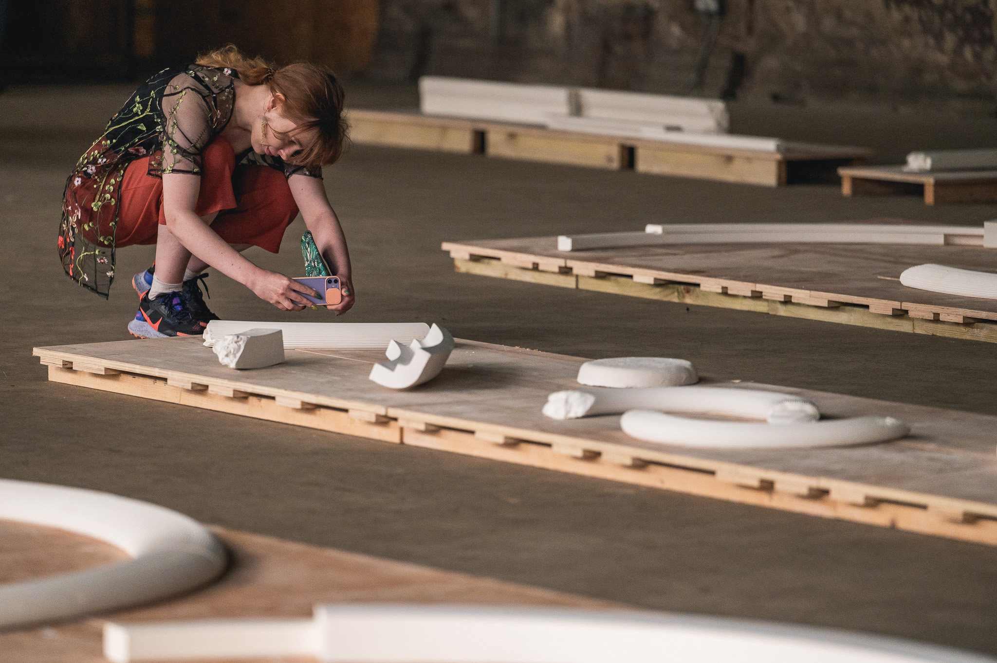 A woman crouches and takes a photo of abstract white sculptures arranged on wooden platforms in a spacious, industrial-style gallery.