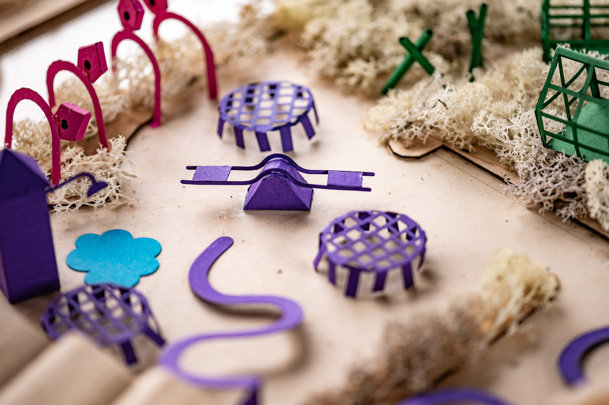 Colourful miniature playground equipment, including a purple seesaw, round structures, and green and pink climbing frames, arranged on a board with patches of artificial grass and tan pathways.