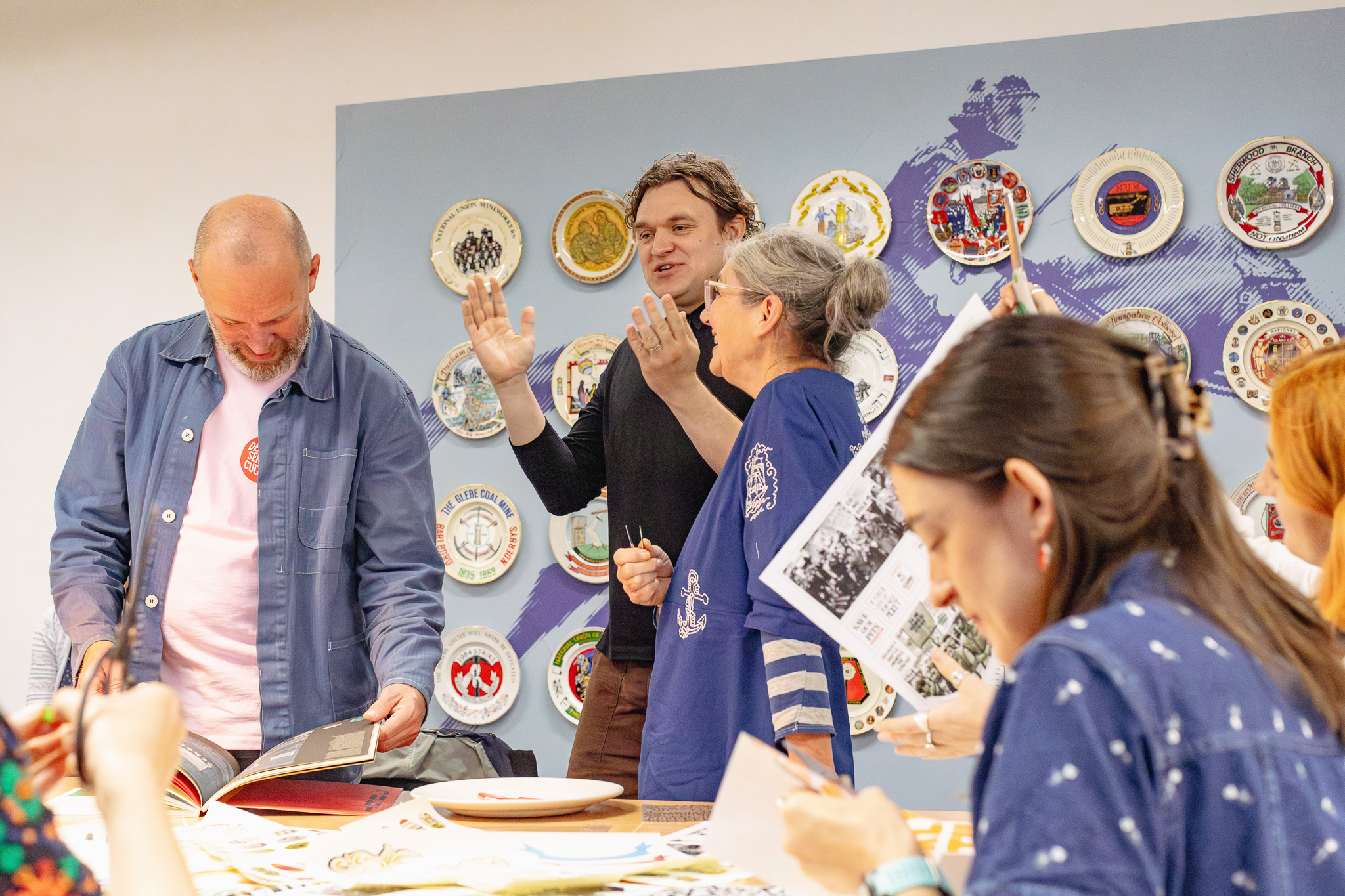 A group of people gather around a table, looking at books and papers, laughing and talking. Decorative mining plates are displayed on the wall behind them.
