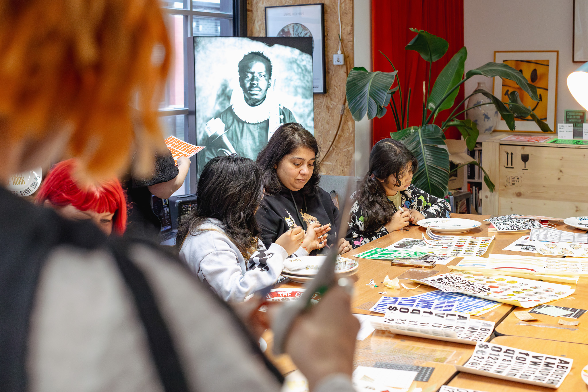A group of people sit around a table, engaged in arts and crafts with various colourful materials. A black and white portrait is displayed in the background, and plants and art supplies decorate the creative, well-lit room.