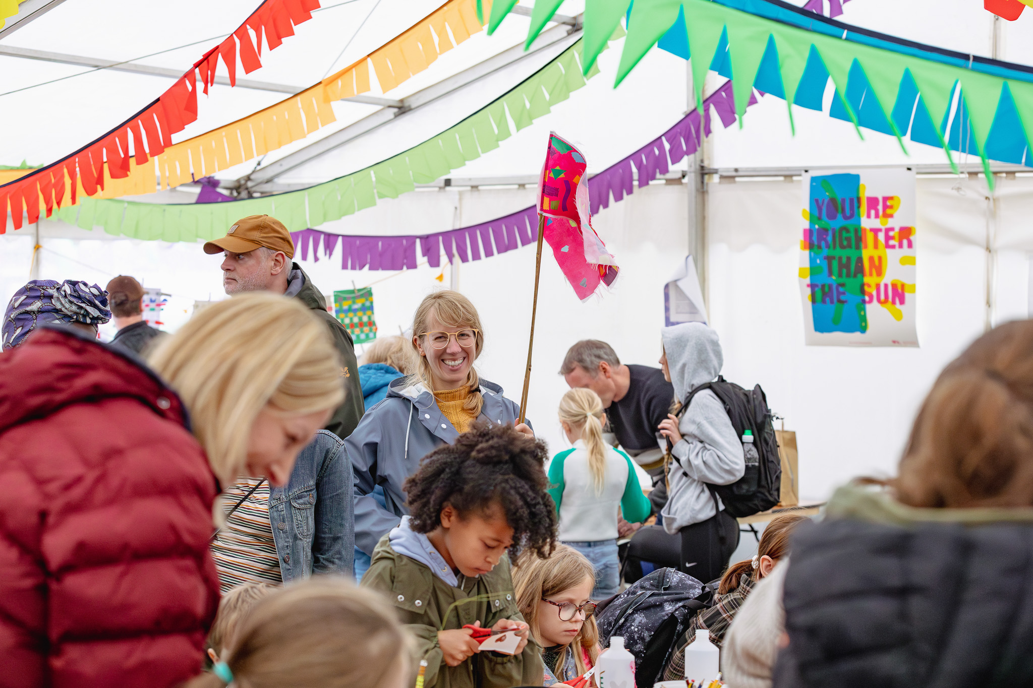A busy, colourful indoor fair with people of all ages, vibrant pennant banners overhead, and a sign reading You’re brighter than the sun on the wall. Children and adults are engaged in arts and crafts activities.
