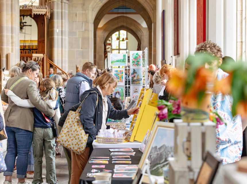 People browse tables with art and crafts at an indoor market or fair inside a large, bright building with high ceilings and arched windows. Some attendees look closely at displays while others converse.