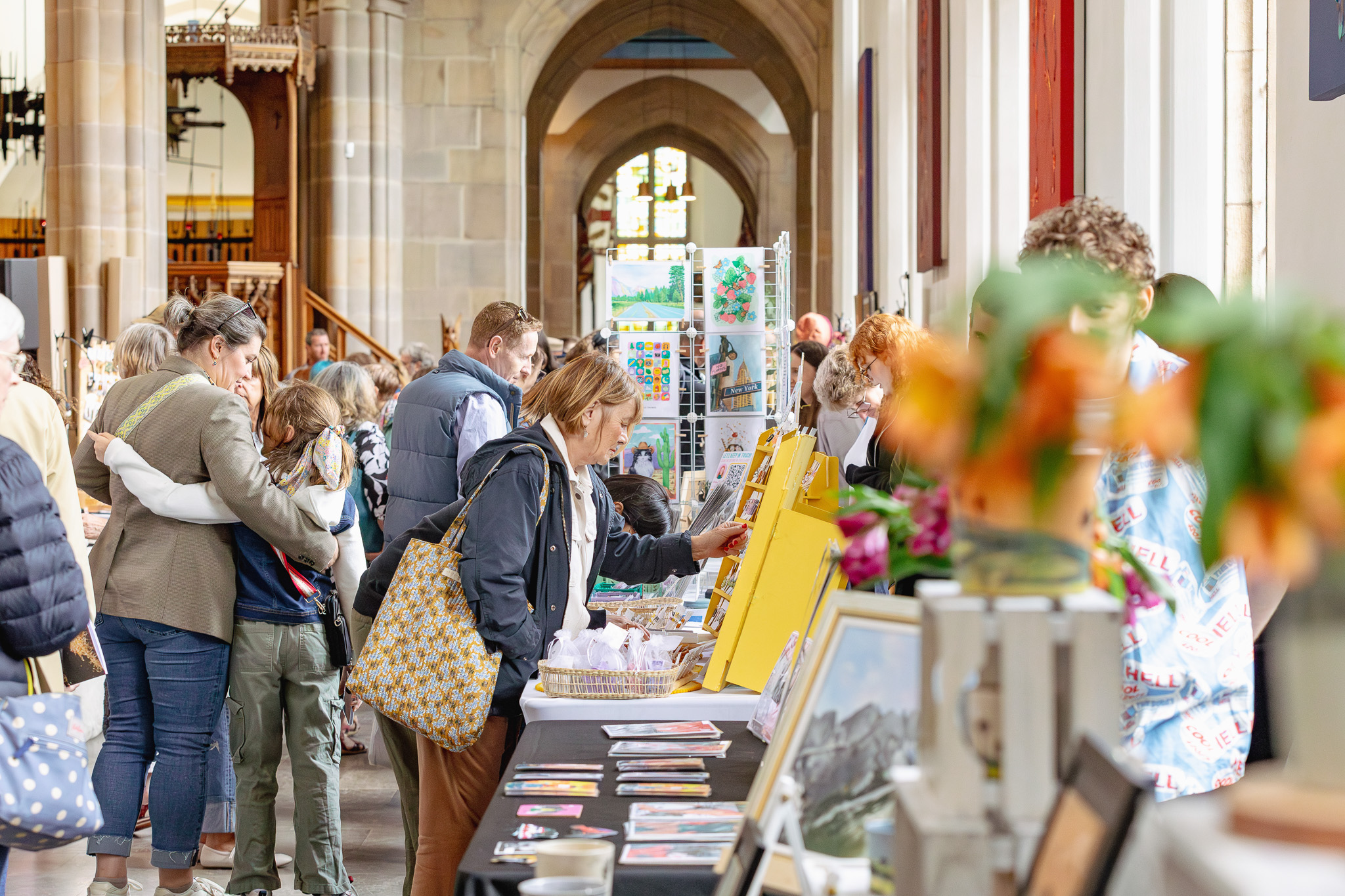 People browse tables with art and crafts at an indoor market or fair inside a large, bright building with high ceilings and arched windows. Some attendees look closely at displays while others converse.