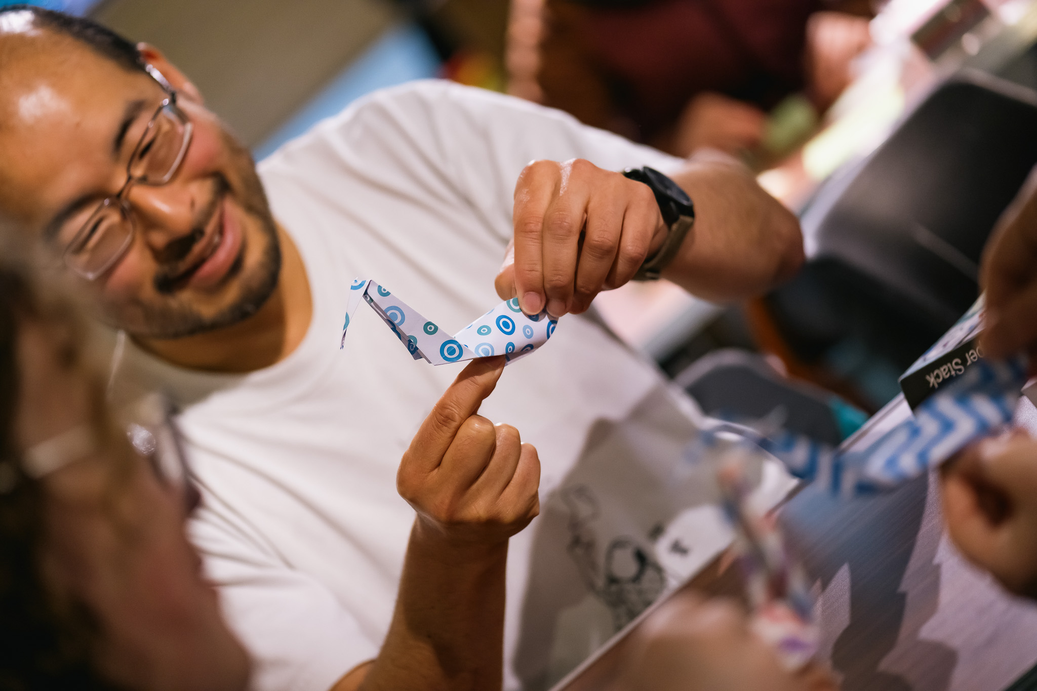 A smiling man wearing glasses and a white shirt holds a patterned paper crane while participating in a workshop.
