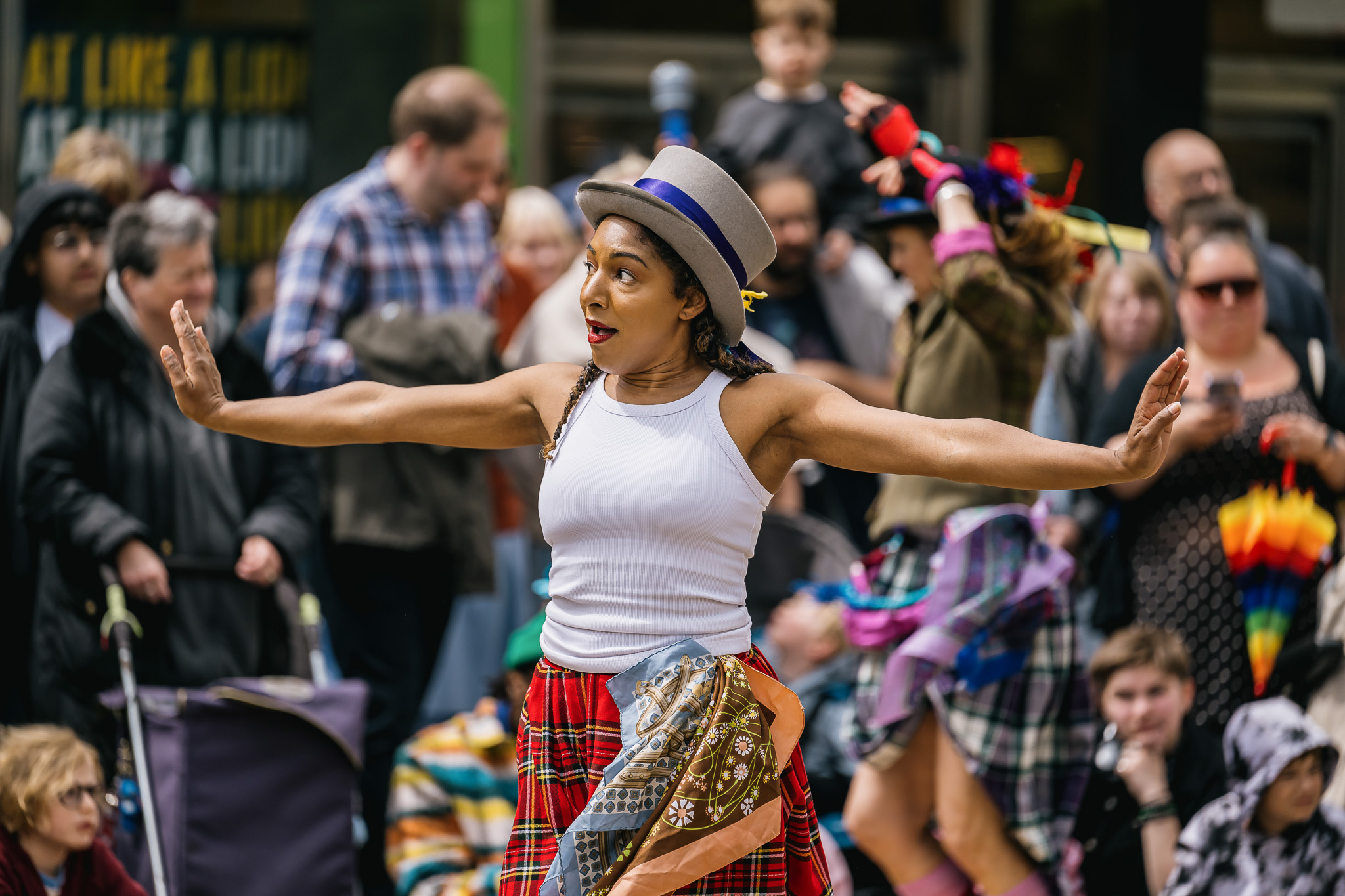 A performer in a white vest, checked skirt, and hat dances with arms outstretched, surrounded by a crowd of onlookers at an outdoor event. The scene is lively and colourful.