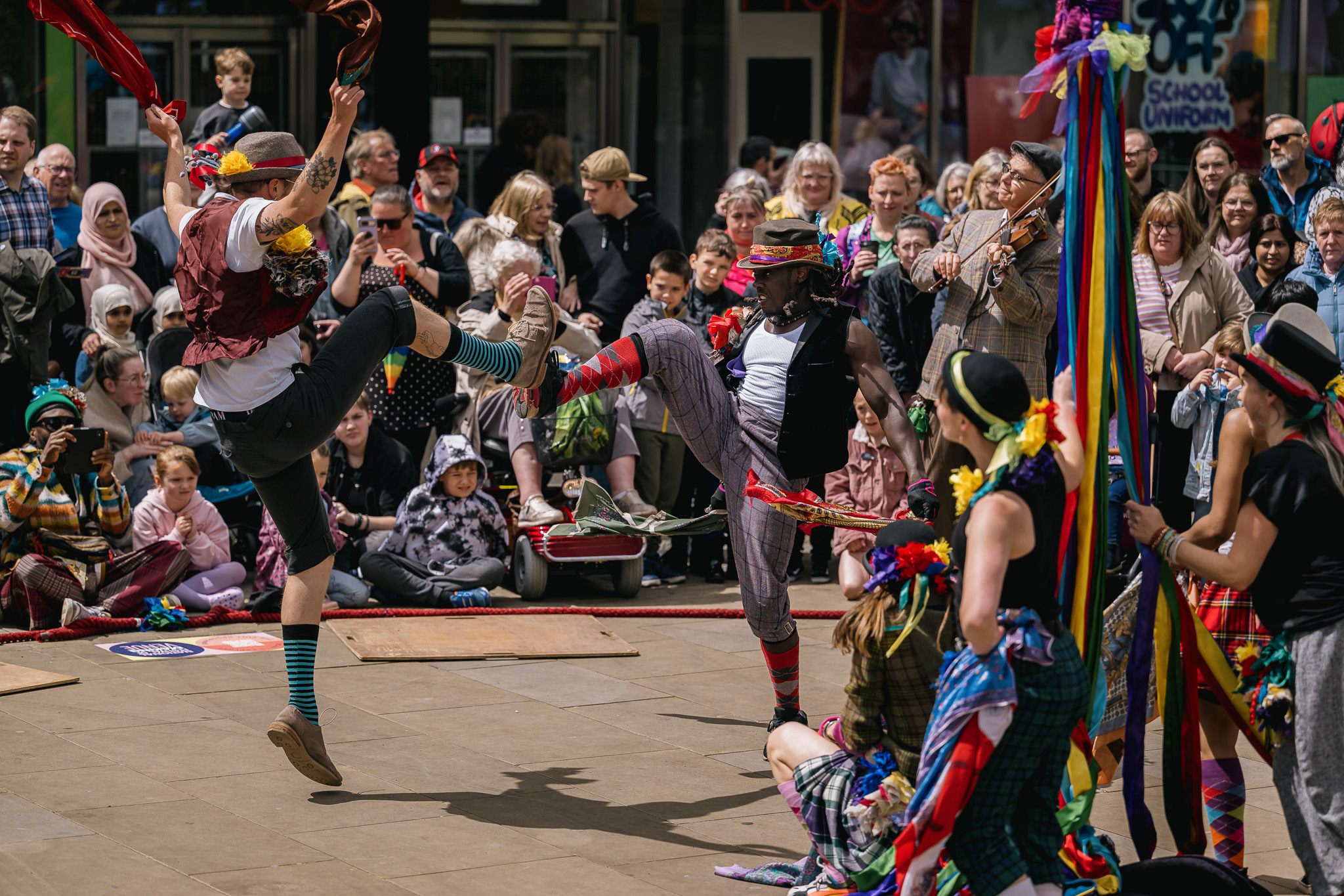 Street performers dressed in colourful, eccentric costumes dance energetically in front of a large, diverse crowd; two dancers are mid-air with high kicks, while onlookers watch and smile.