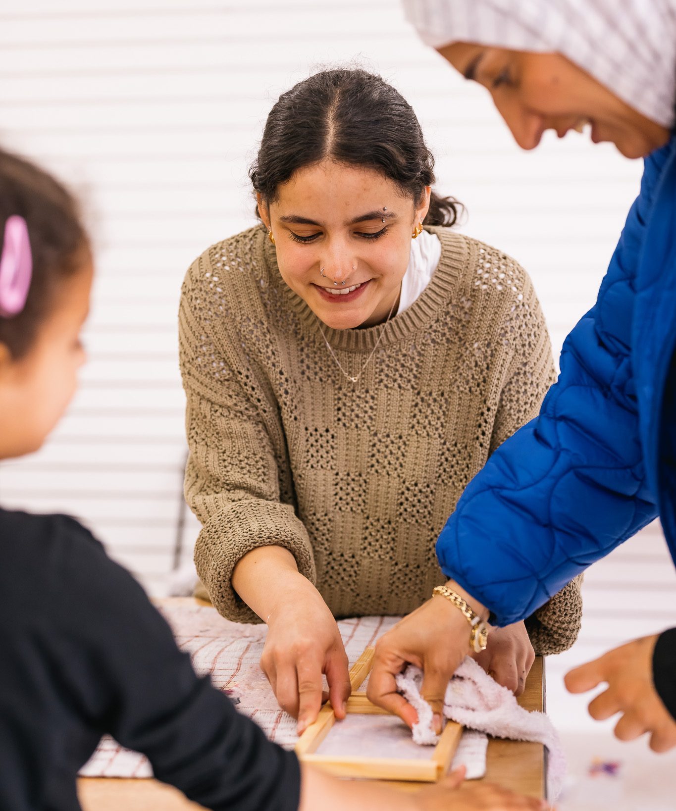 Three people make paper at a workshop. One woman in a brown jumper smiles as she helps hold a wooden frame, whilst two others assist, reaching in from the sides.