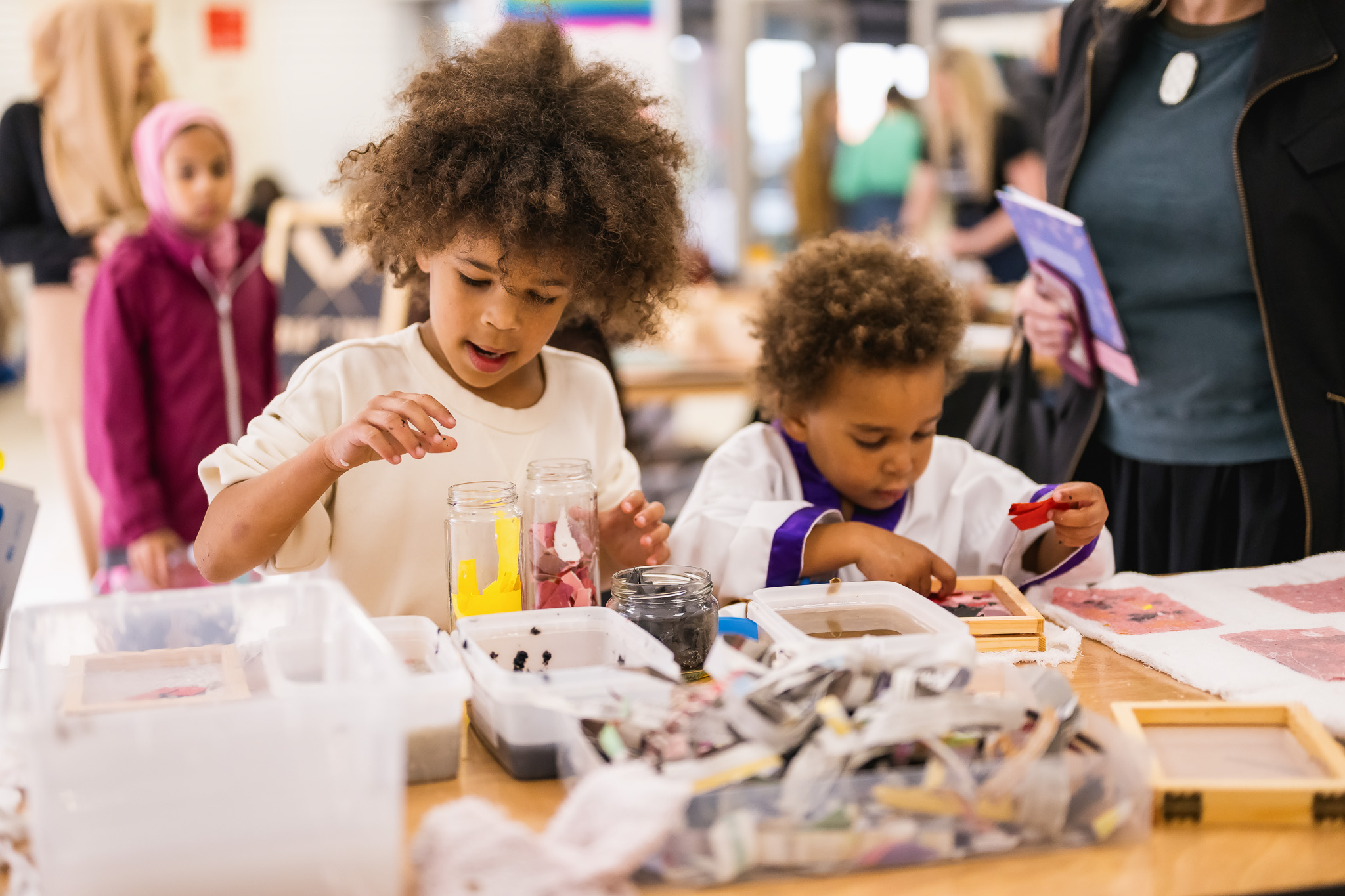 Two young children sit at a table covered with art supplies, focusing intently as they work on craft projects in a bright, busy indoor setting with adults and children in the background.